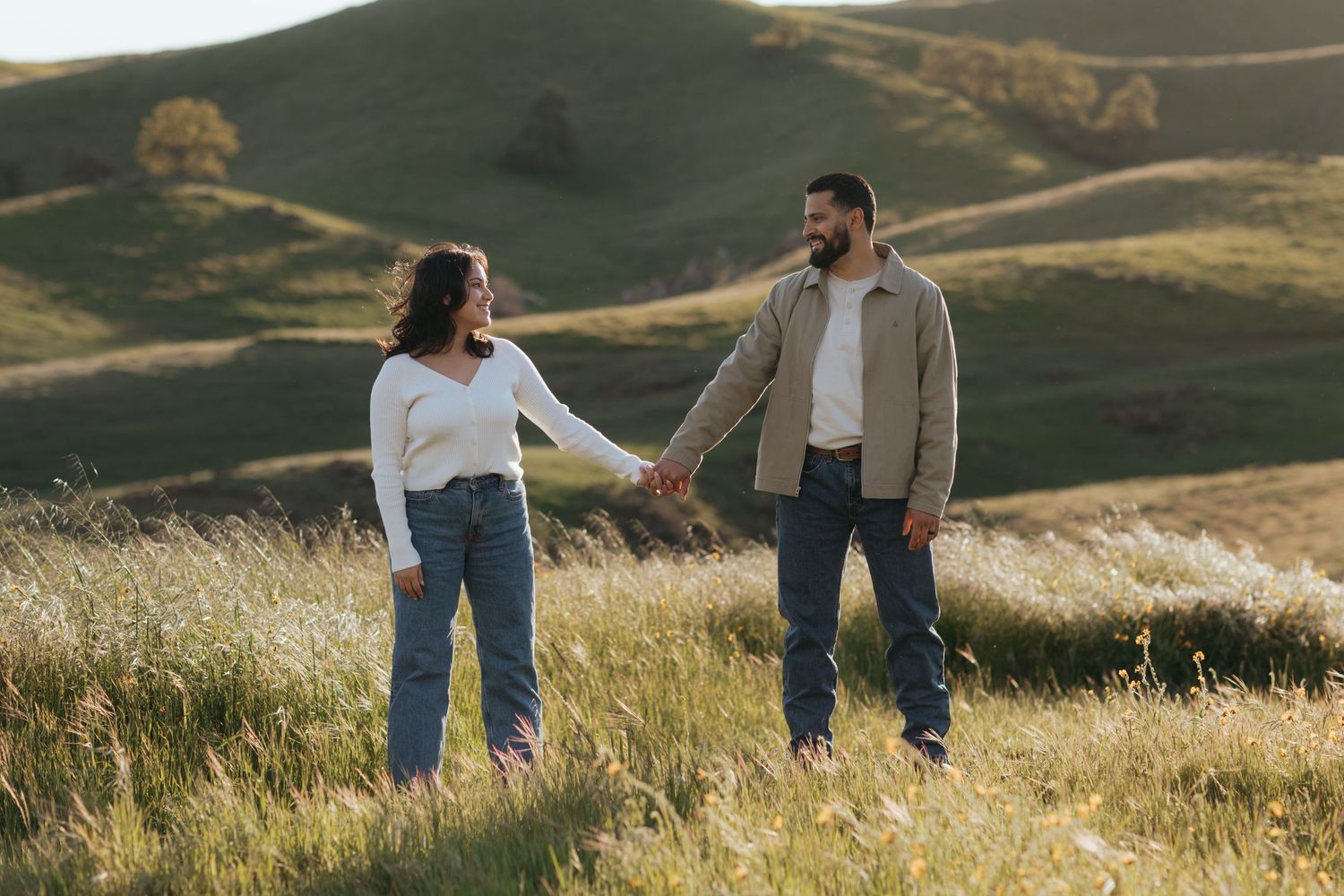 man and woman standing apart, holding hands while looking lovingly at each other while also standing on a hilltop of wildflowers.