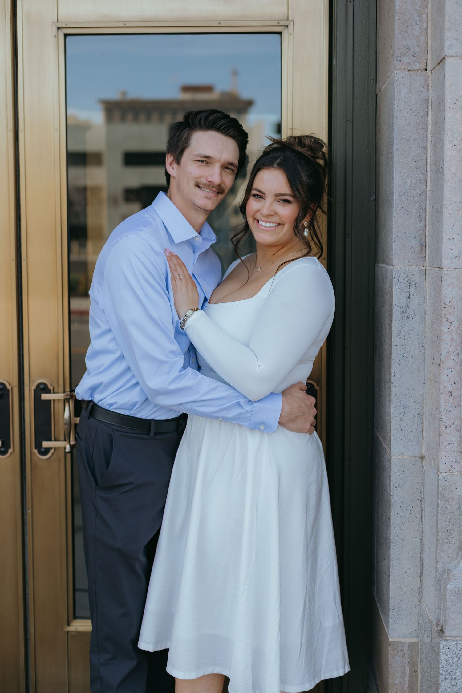 A smiling couple holding each other in front of a golden framed glass door.