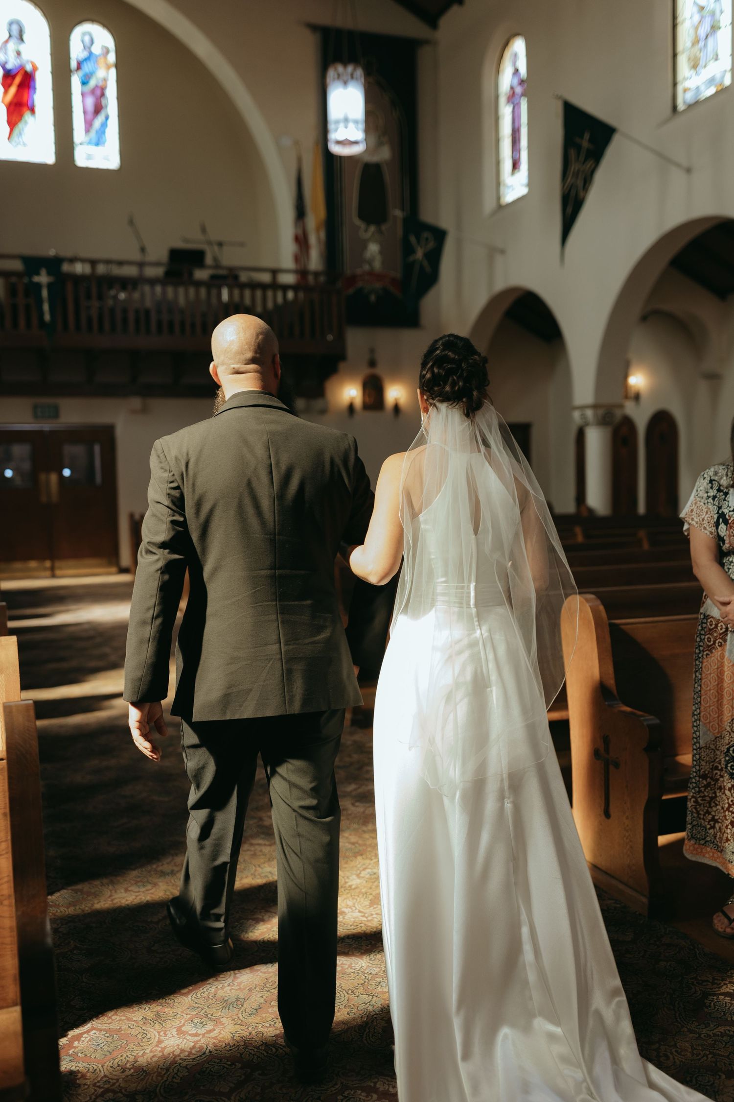 A wedding couple with backs facing the camera walking down the aisle in a Catholic church with stained glass windows.
