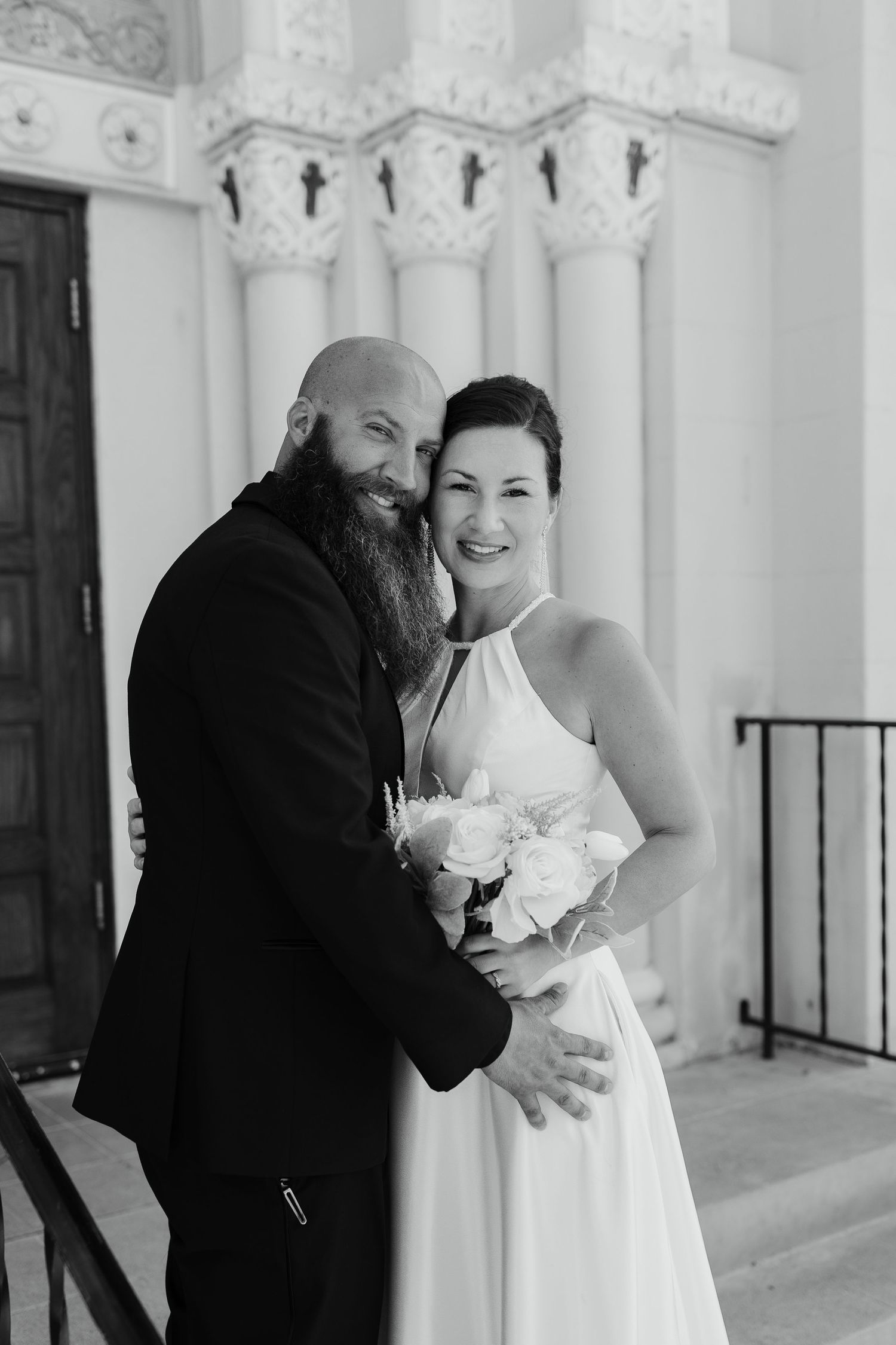 black and white image of a wedding couple smiling at the camera in front of a church
