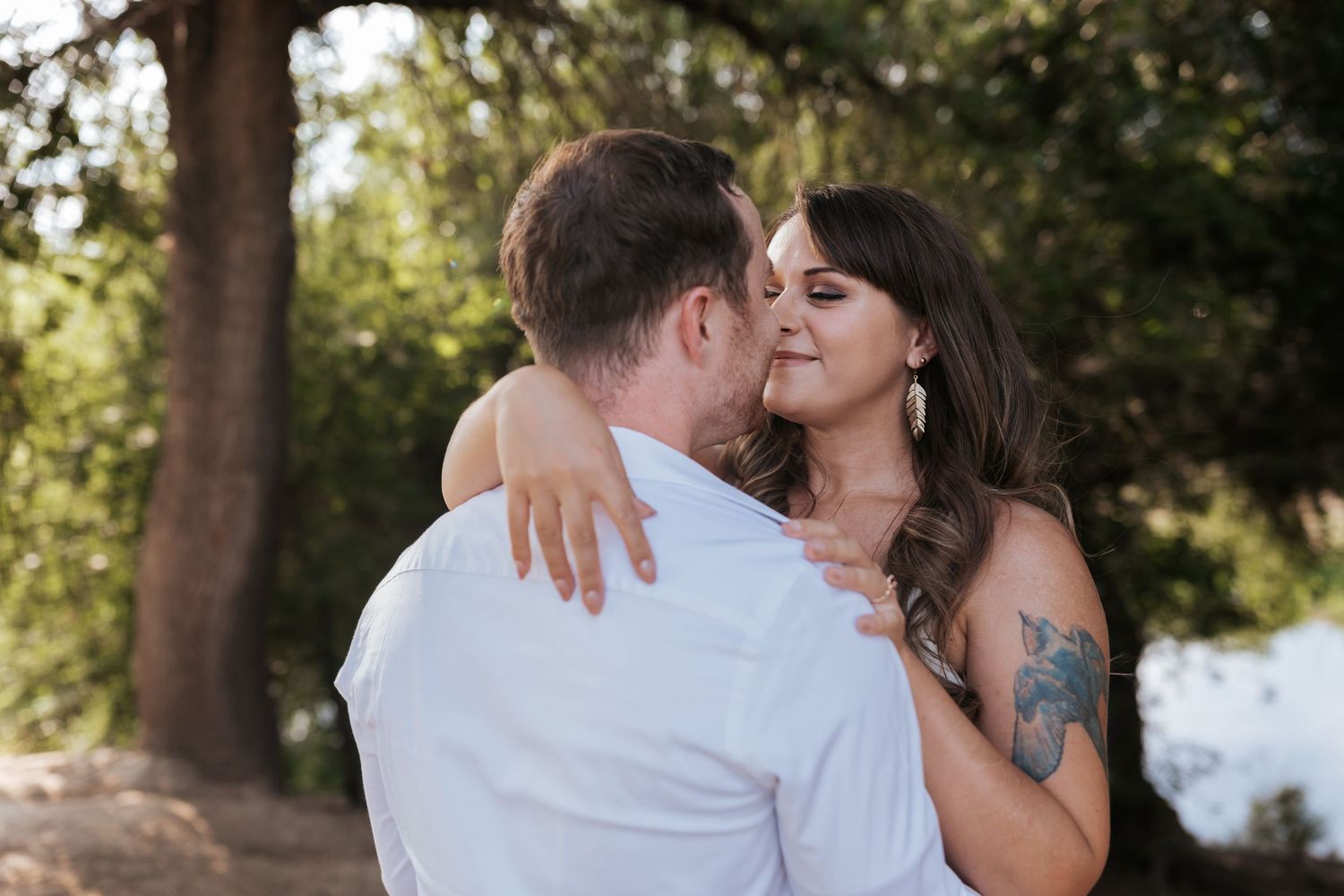 Couple embracing outdoors under trees, standing close in warm natural light during an intimate moment.