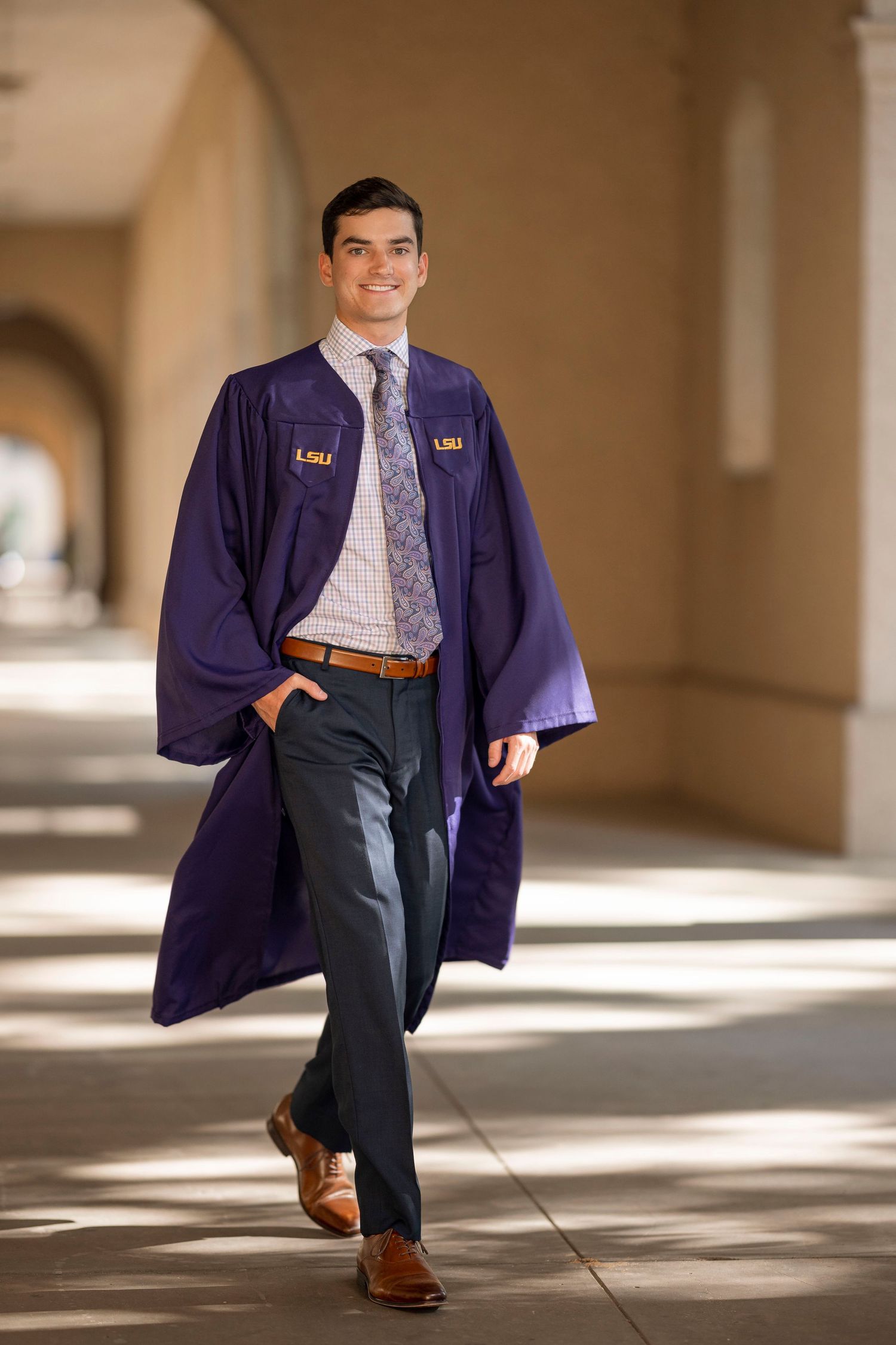 LSU graduate in navy graduation gown and business attire walks confidently through campus archway.