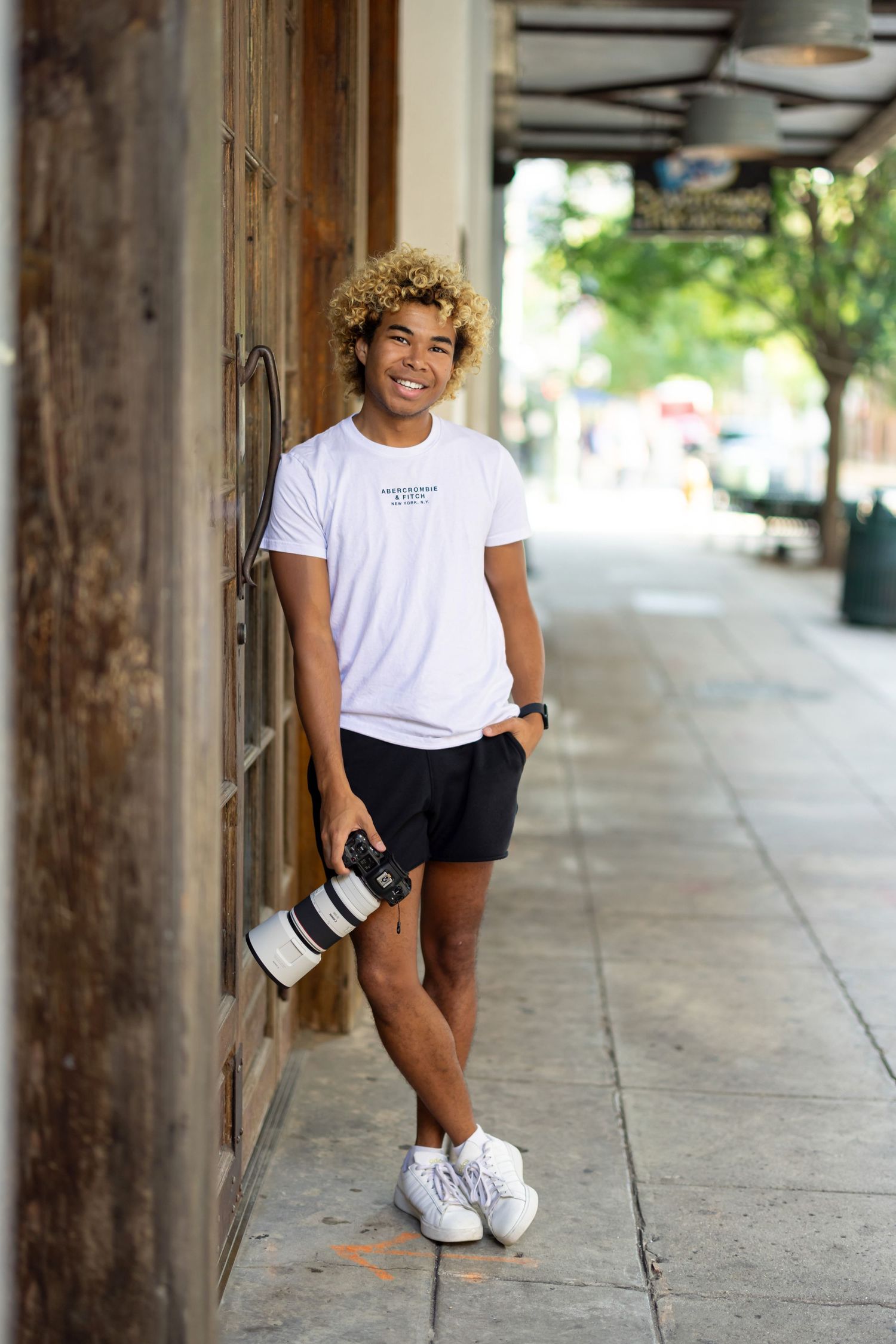 Photographer in casual white t-shirt and shorts leans against wall with professional camera gear.