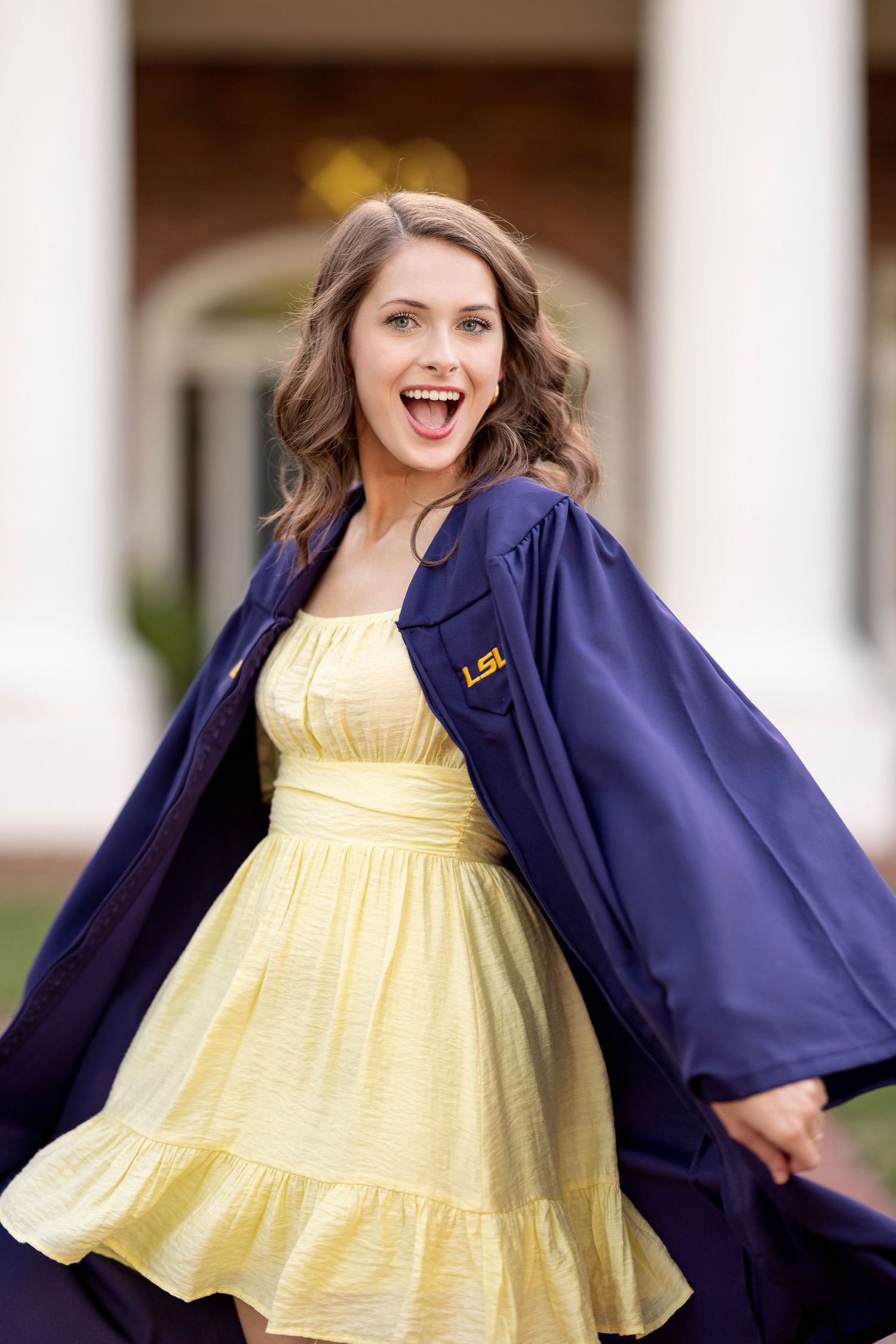 Graduate in navy gown and yellow sundress poses joyfully on campus during graduation photoshoot at LSU.