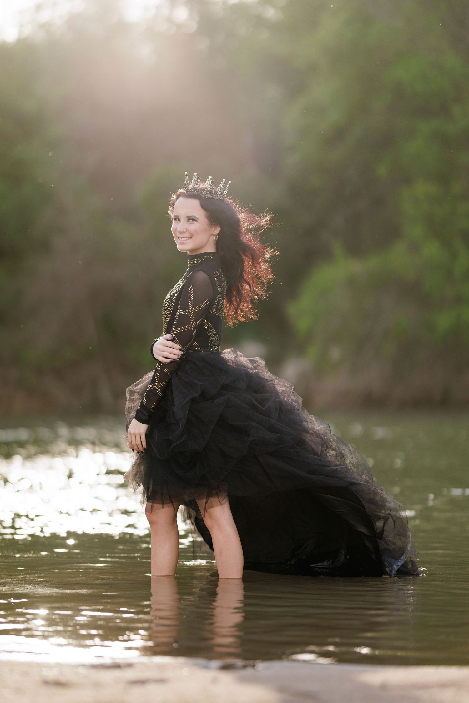 Model in dramatic black tulle dress poses in water during sunset photoshoot.