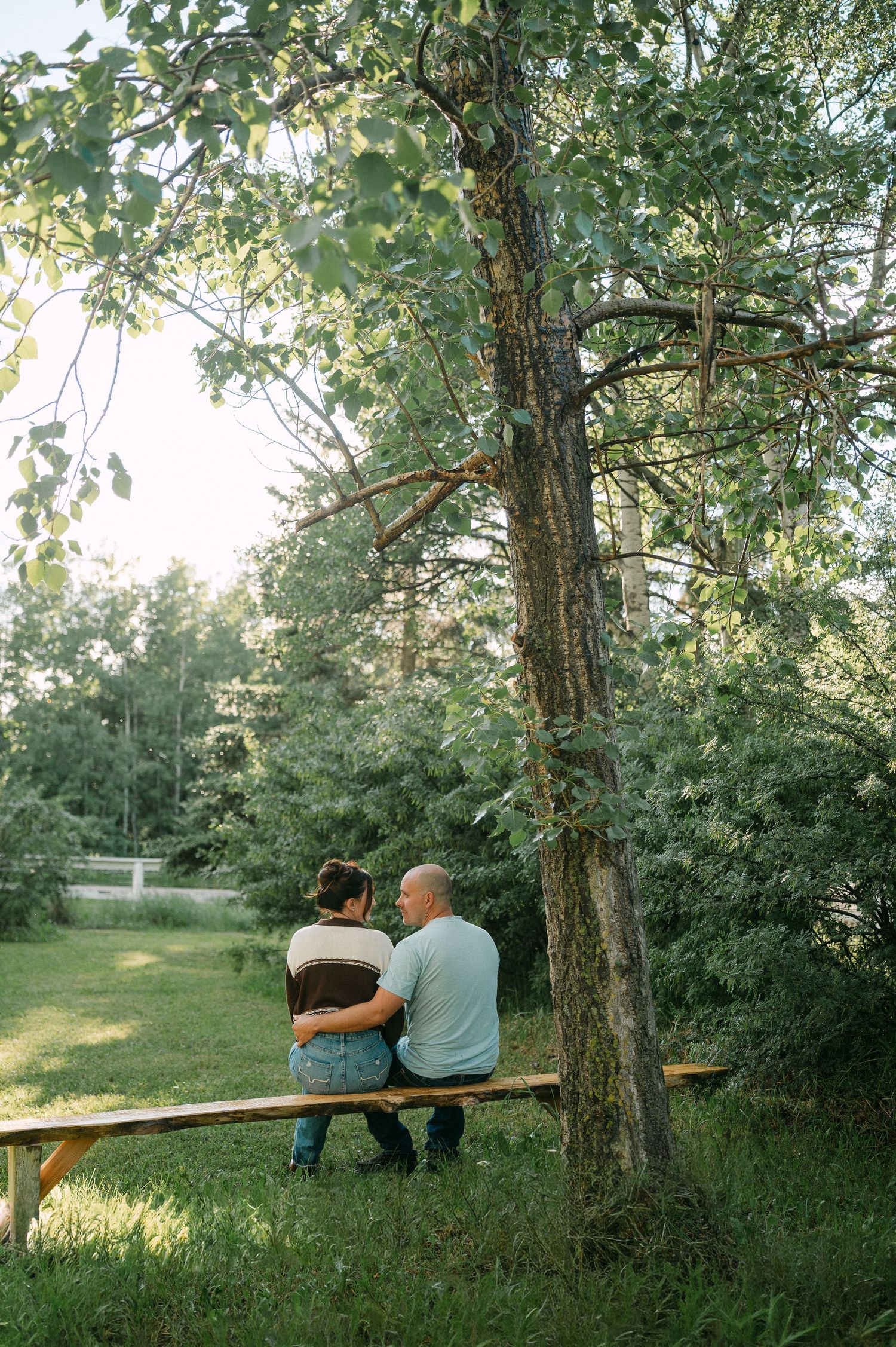 Couple sitting close together on a wooden bench under a tree in a lush green park setting.