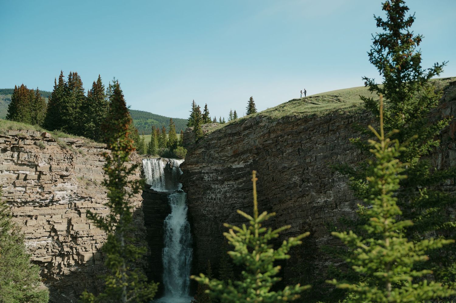 A stunning waterfall cascades down rocky cliffs surrounded by evergreen trees. Couple in the distance, engagement session.