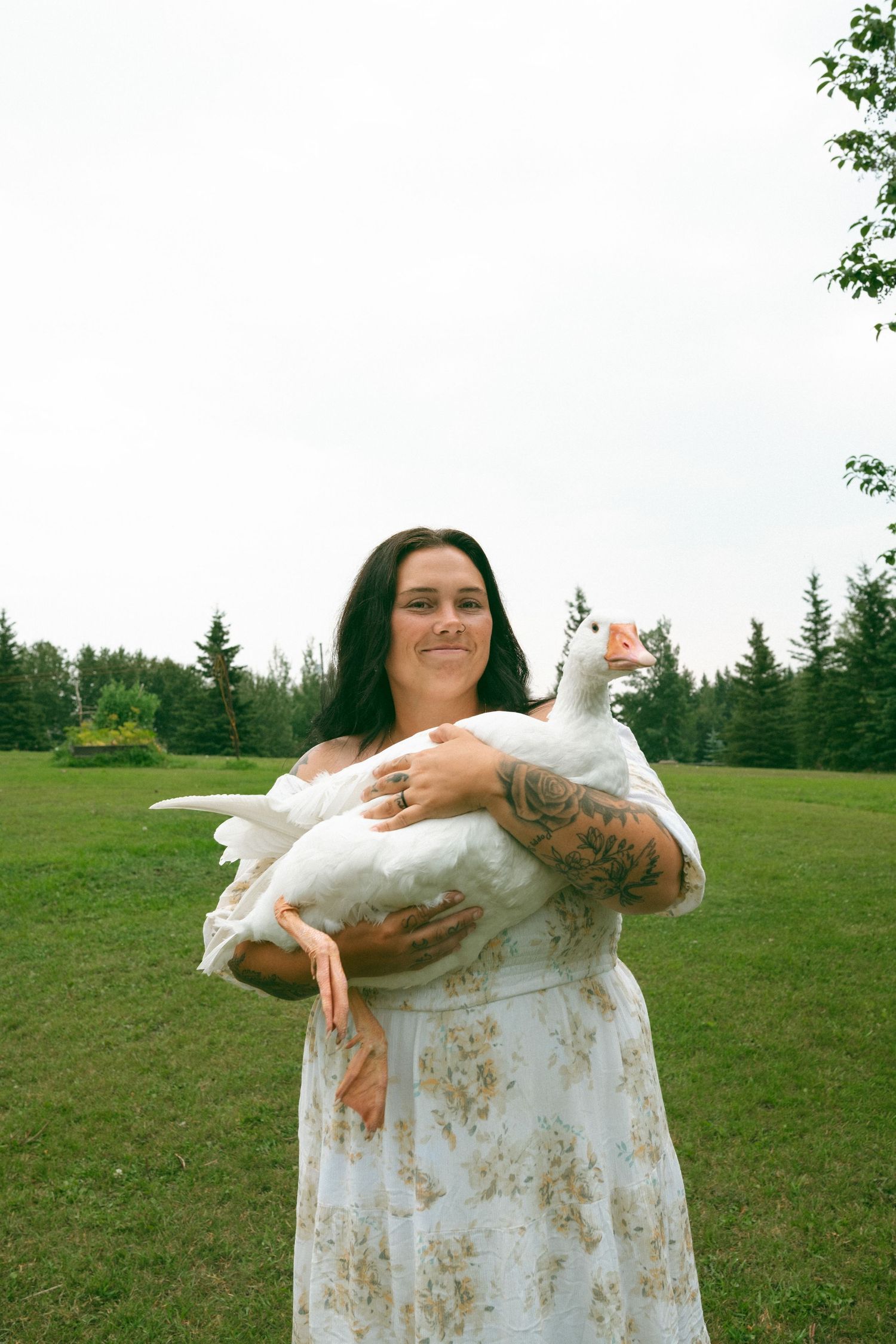 Woman in floral dress holding a white goose outdoors on green grass with trees in the background.
