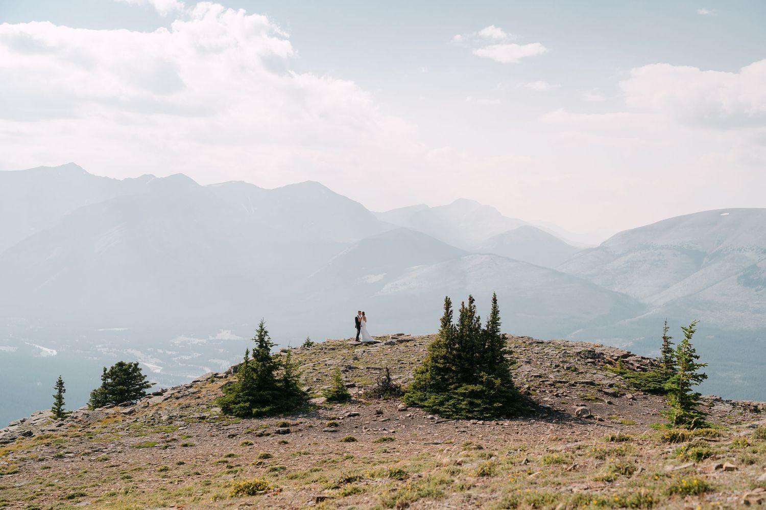 A lone hiker stands on a rocky mountain summit surrounded by evergreen trees with sweeping mountain views.