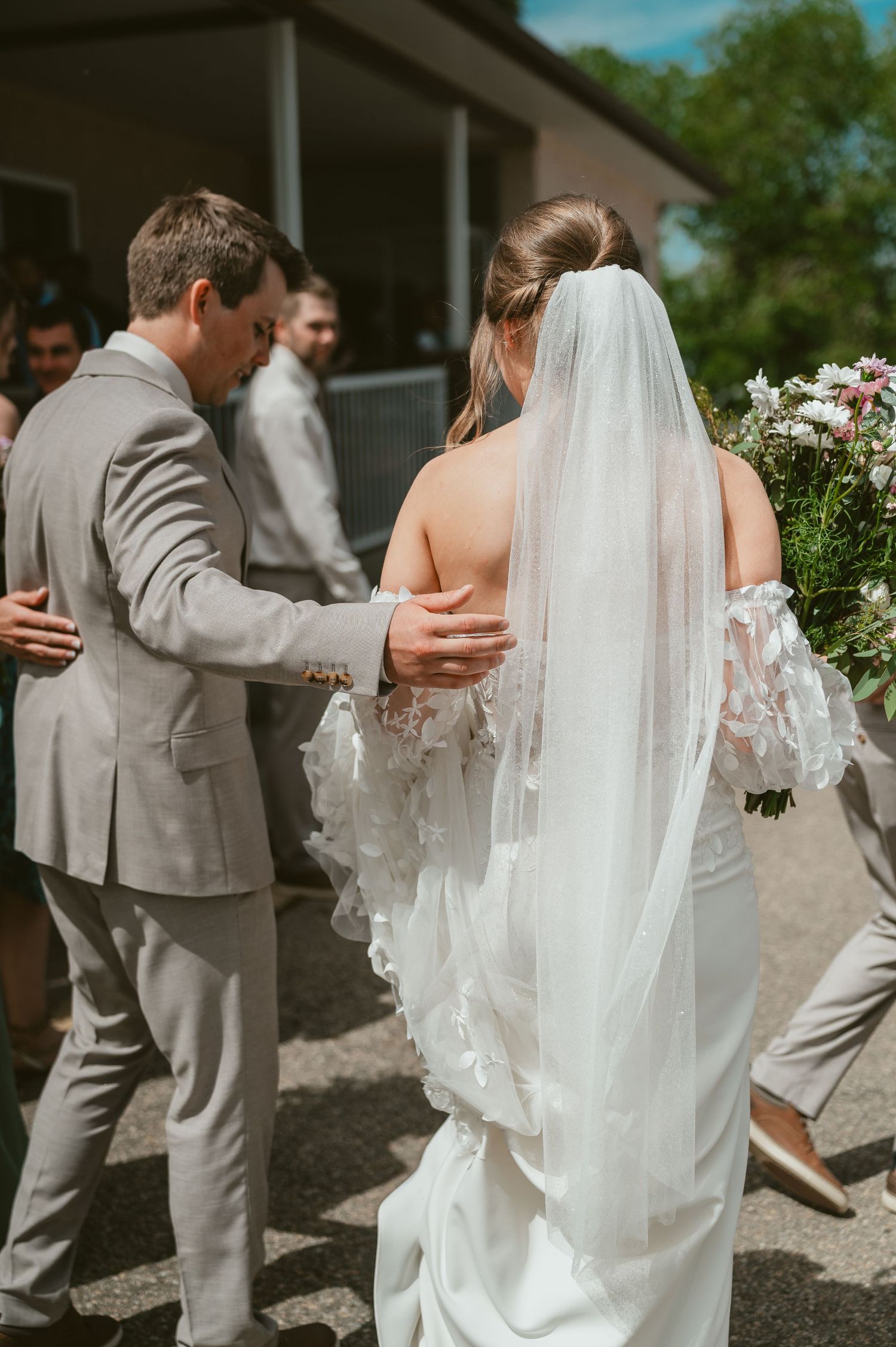 Bride in white gown and veil walks with groom in gray suit at outdoor wedding ceremony.