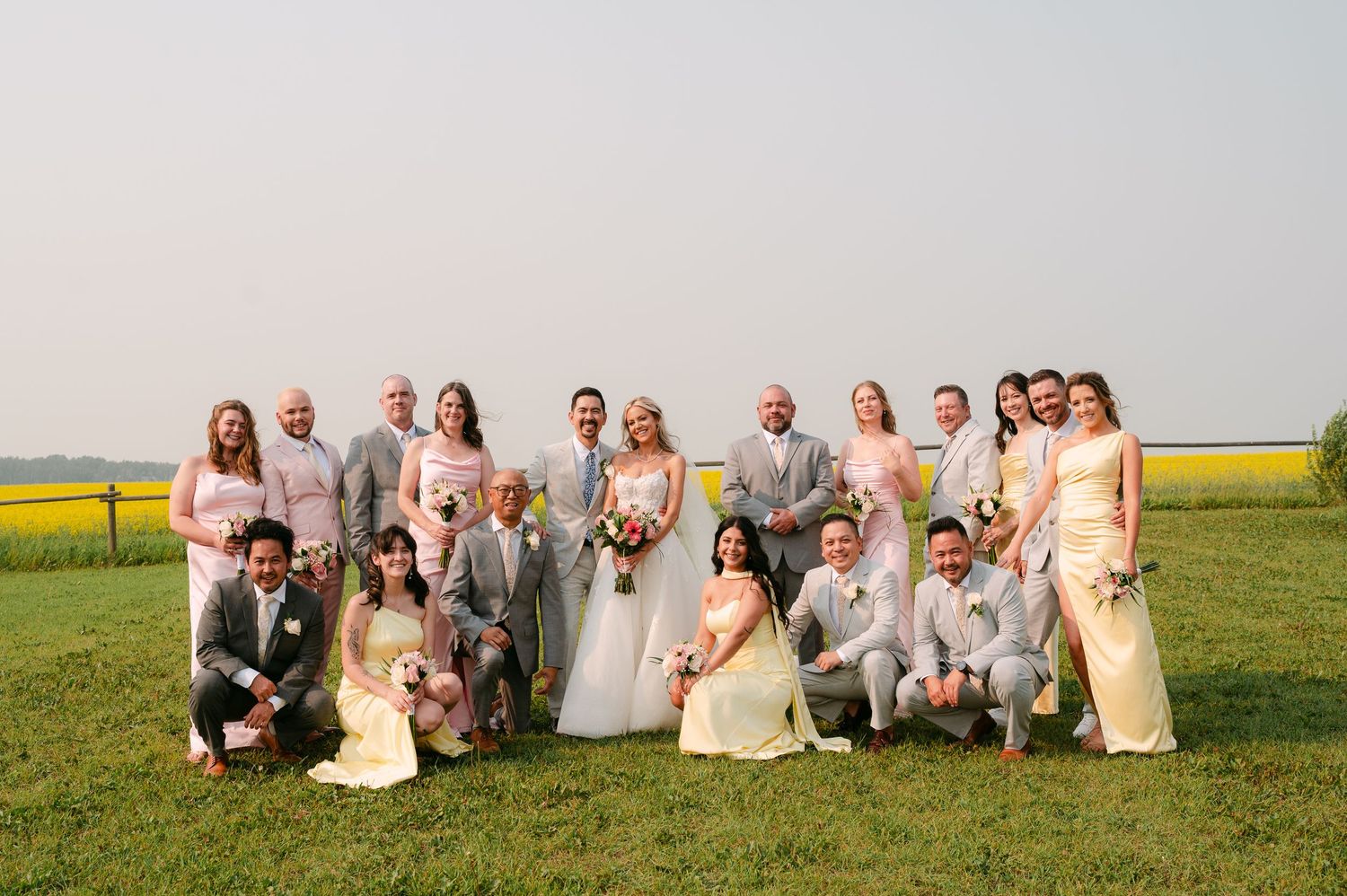 Large wedding party posing outdoors in a green field, bridesmaids in pink, groomsmen in gray suits, bride in white gown.