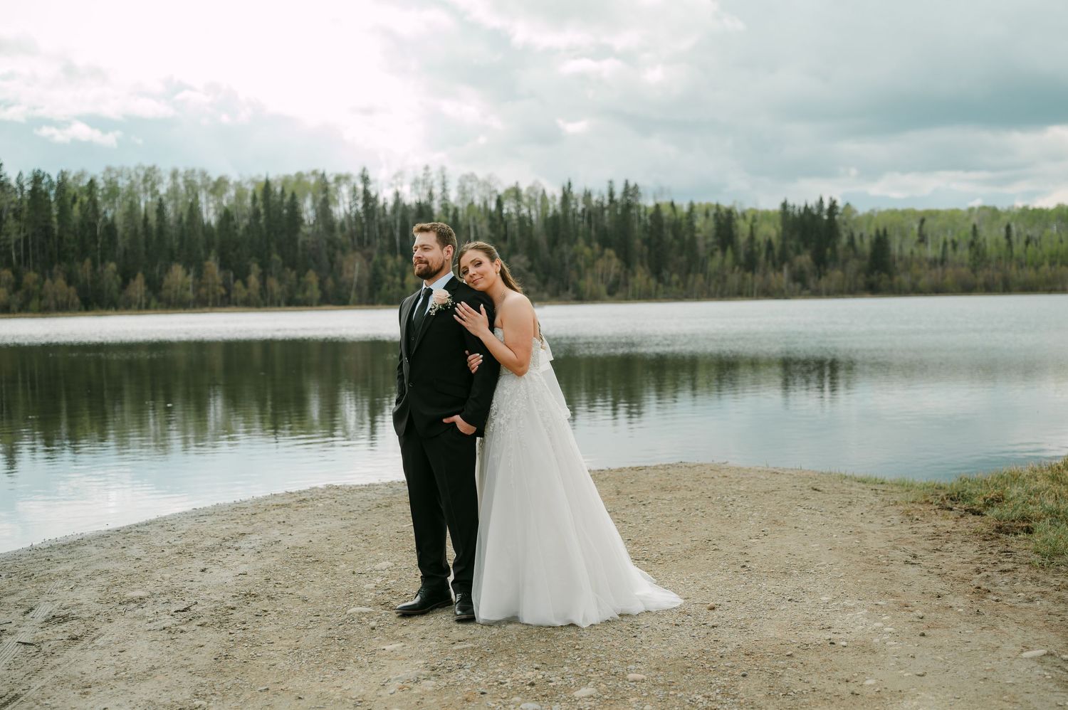 Bride and groom posing together by a serene lake surrounded by evergreen trees on their wedding day.