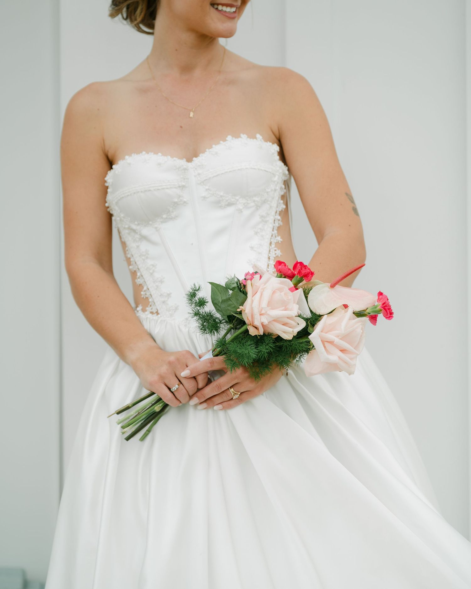 Bride in strapless sweetheart corset wedding gown holds a small bouquet of blush and pink roses with greenery.