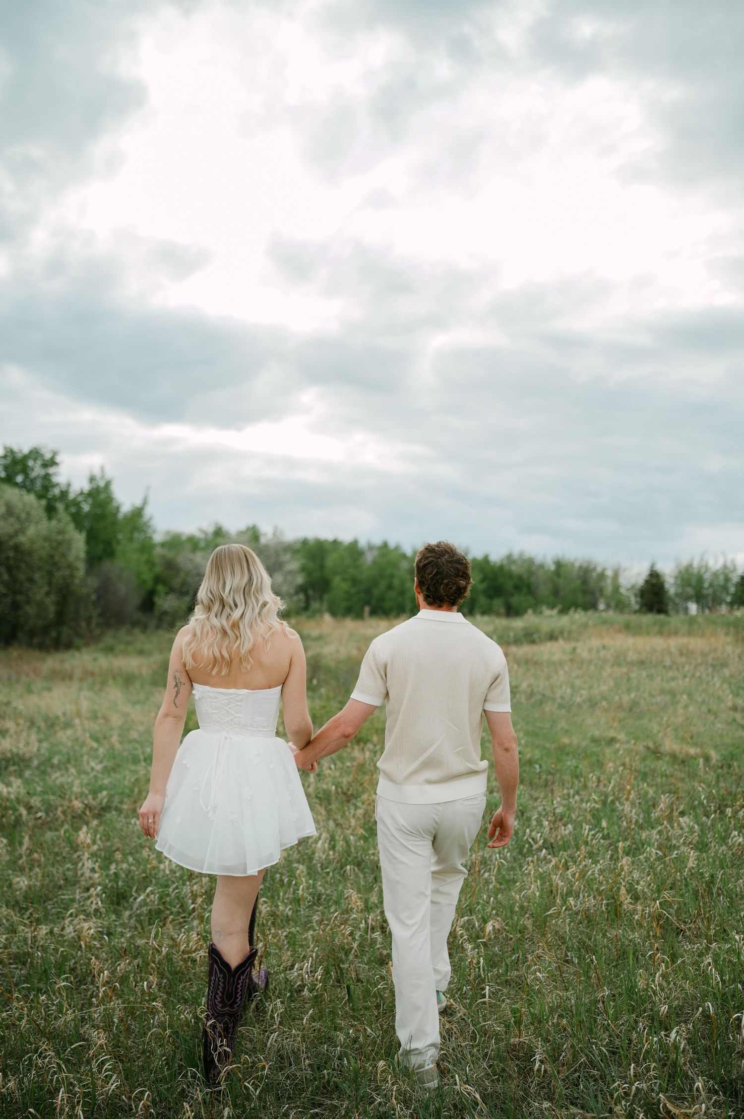 Couple holding hands walking through a green meadow, woman in white dress with cowboy boots. Engaged.