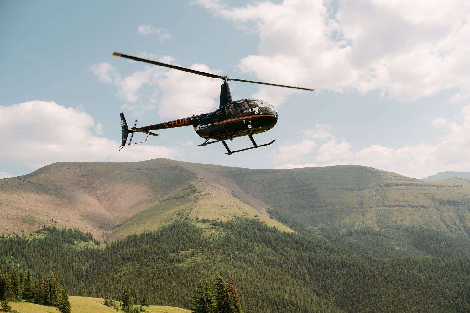 Black helicopter flying over lush green mountain forest landscape under partly cloudy blue sky.