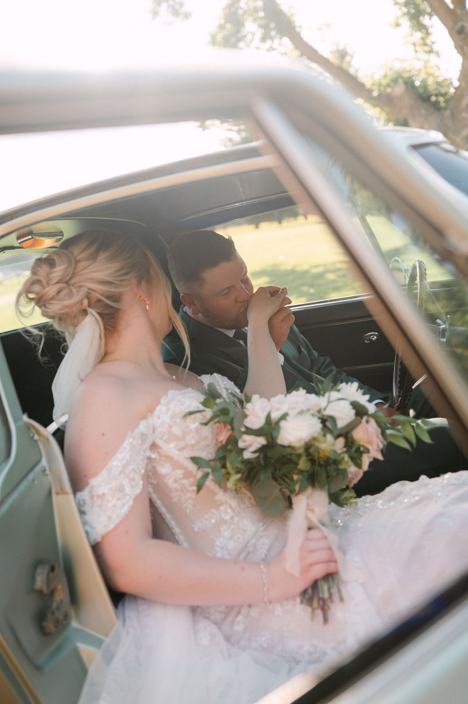 Bride and groom share a romantic moment in a vintage car, bride holding a white and pink floral bouquet.