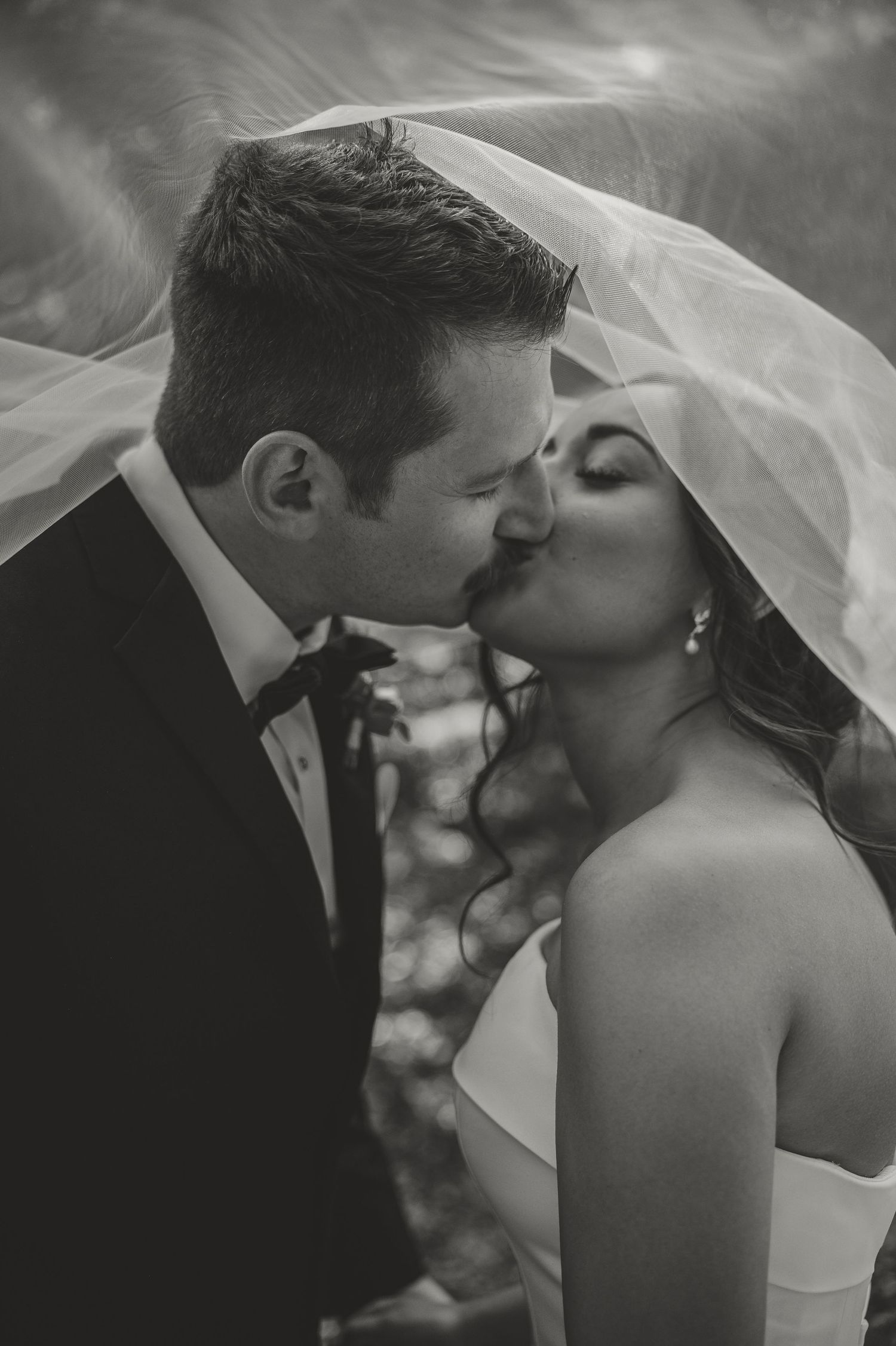 Black and white romantic kiss of bride and groom under flowing veil, outdoor wedding portrait.