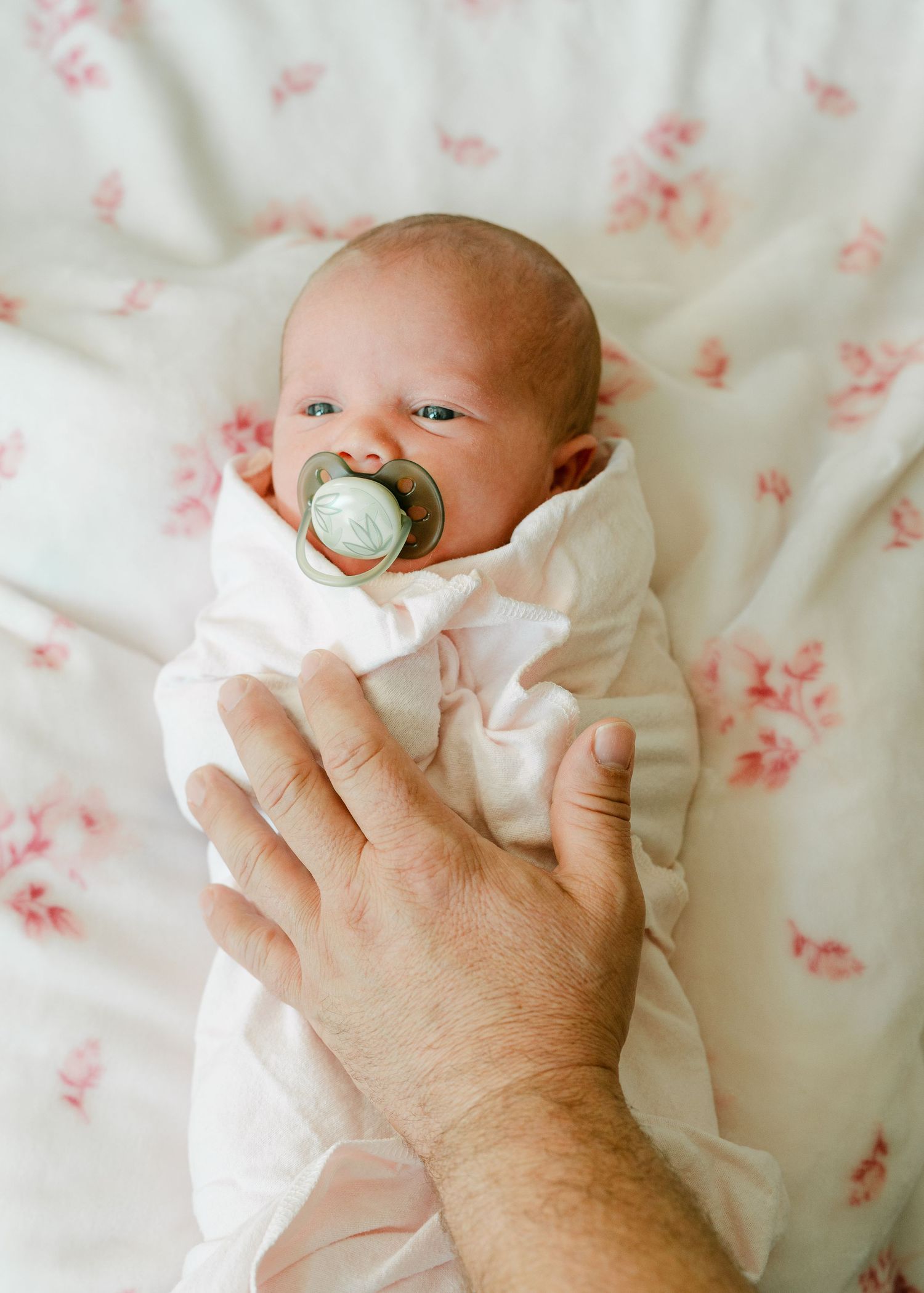 Newborn baby swaddled in white with a pacifier, gently touched by an adult hand on a floral blanket. Lifestyle session.