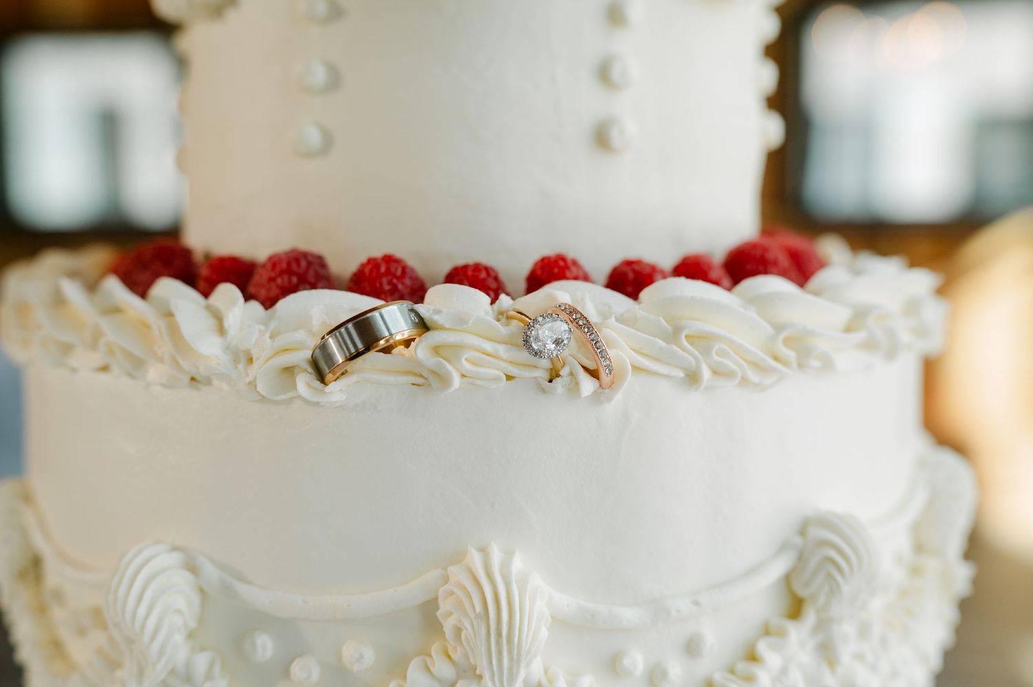 Two gold wedding rings rest on a white tiered wedding cake decorated with fresh raspberries and cream rosettes.