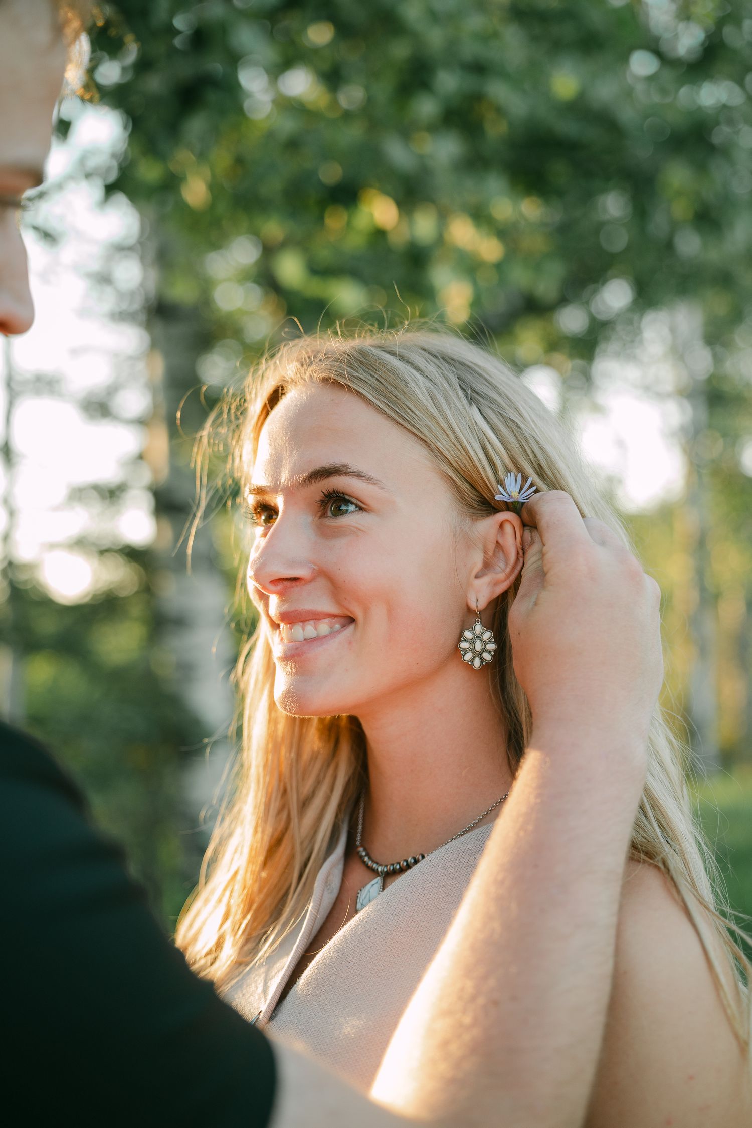 Smiling blonde woman with floral earrings and necklace in golden sunlight outdoors.