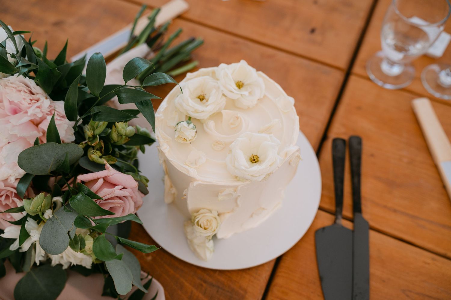 Single-tier white wedding cake with delicate white flowers on a wooden table beside a pink floral bouquet and cake server.