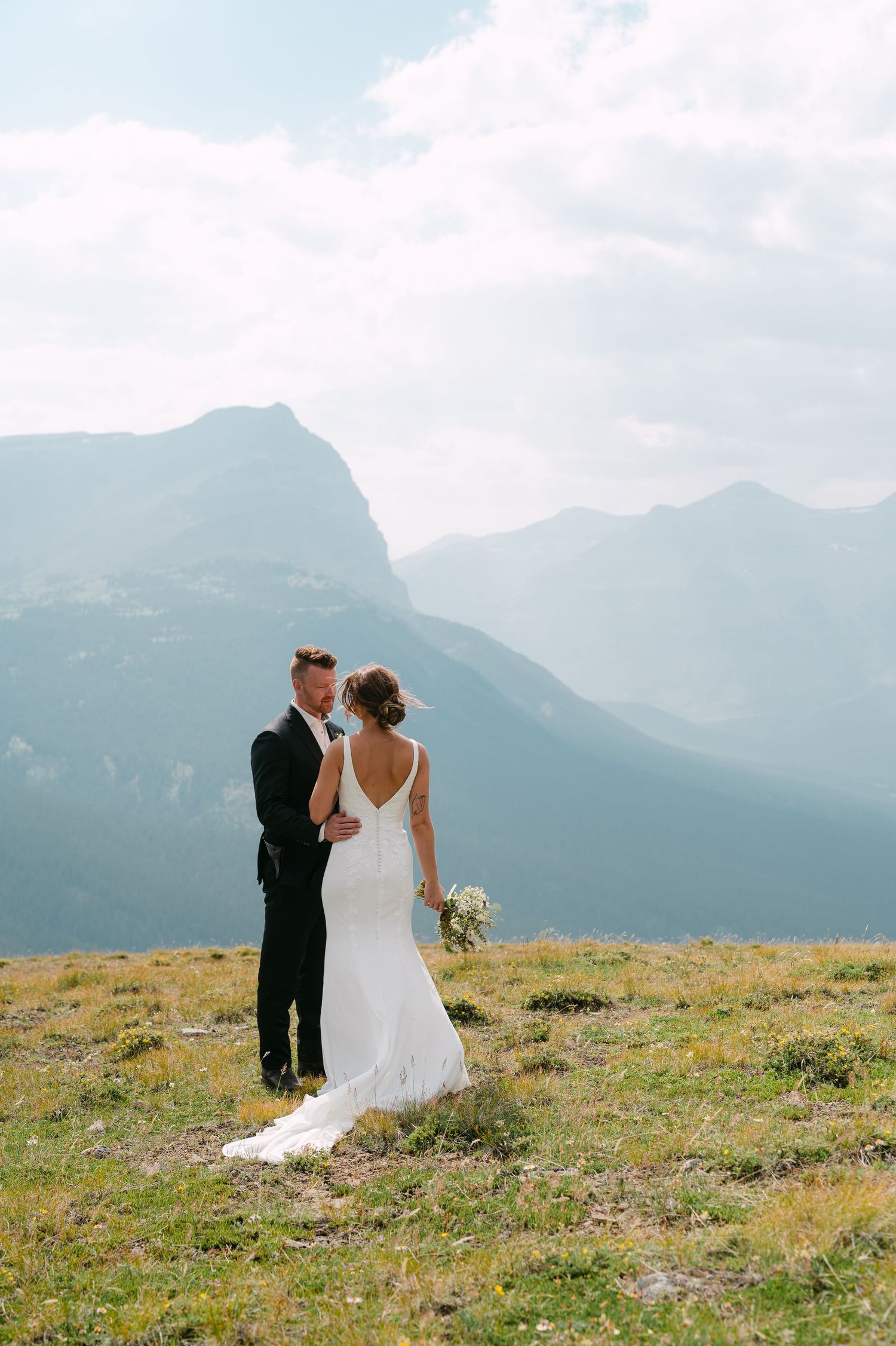 Bride and groom embrace on a mountain summit, with stunning rocky peaks and vast sky backdrop on their wedding day.