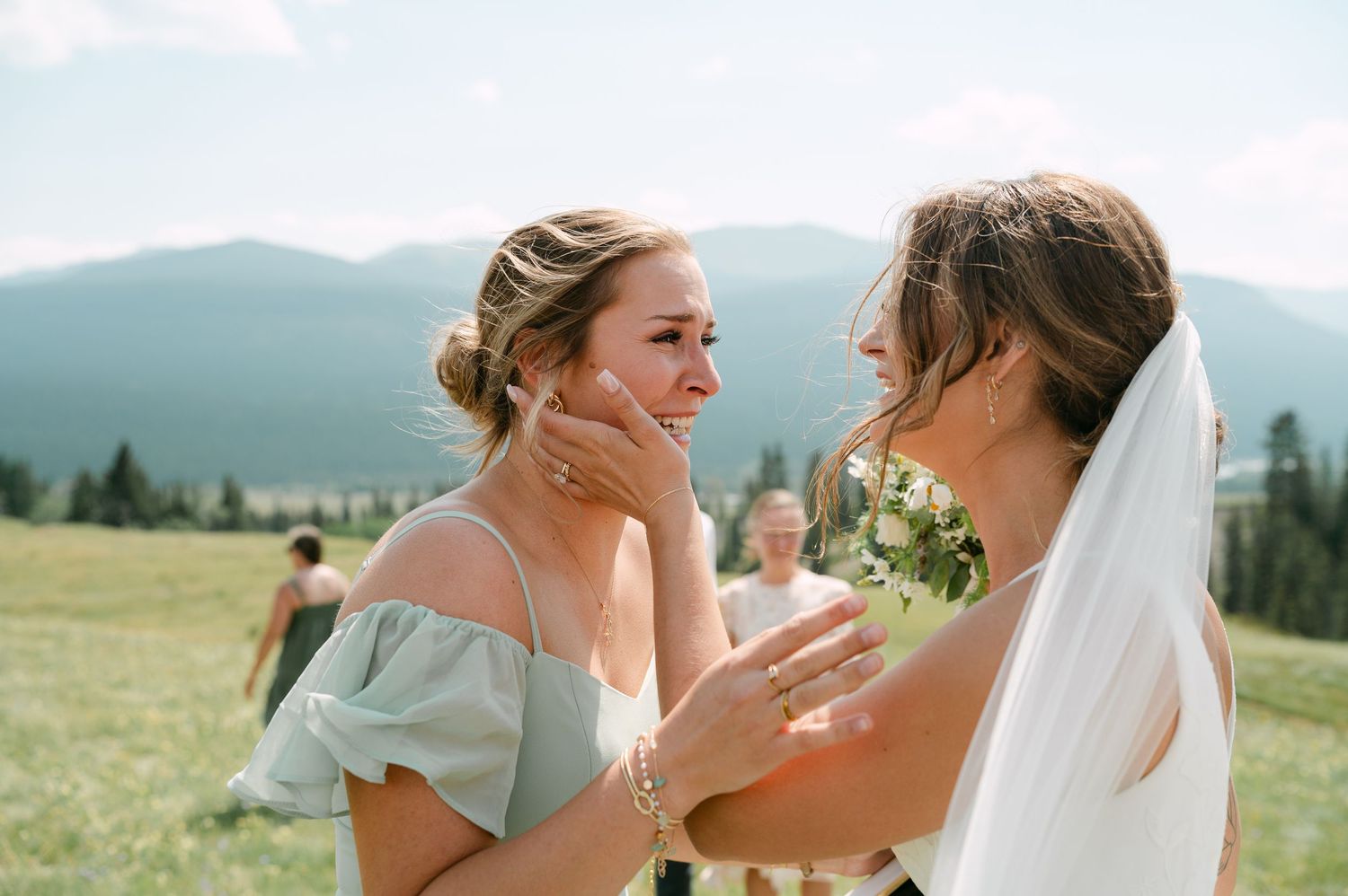 Emotional bridesmaid wiping tears while embracing bride outdoors with mountain backdrop at wedding celebration.