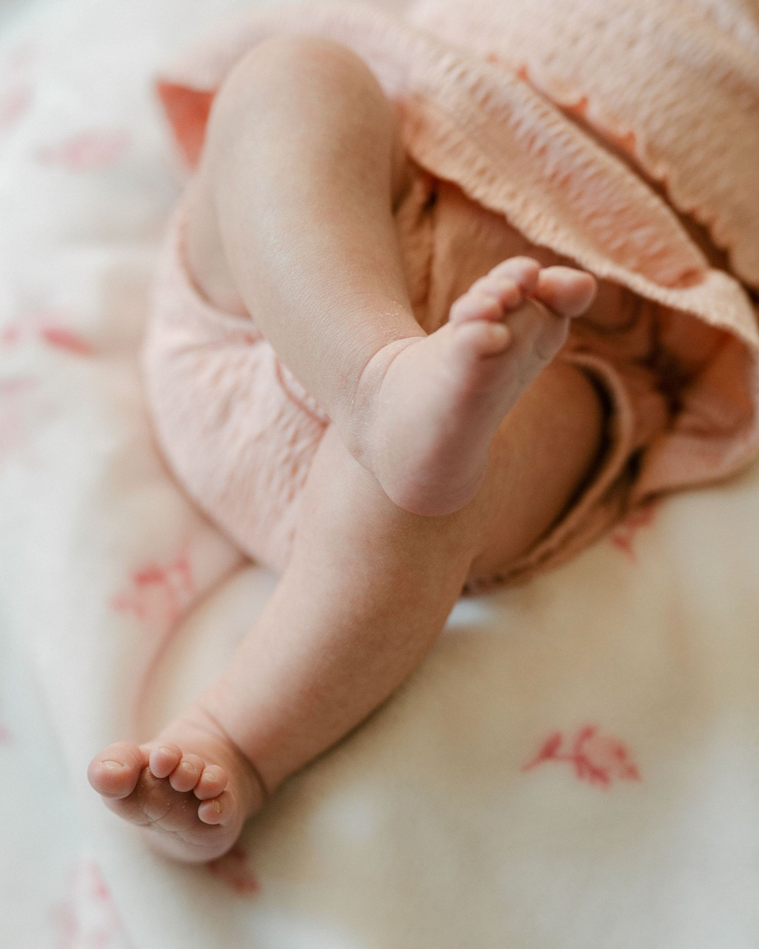 Close-up of a newborn baby's tiny feet and legs resting on a floral blanket in soft pink tones. Detail photos.