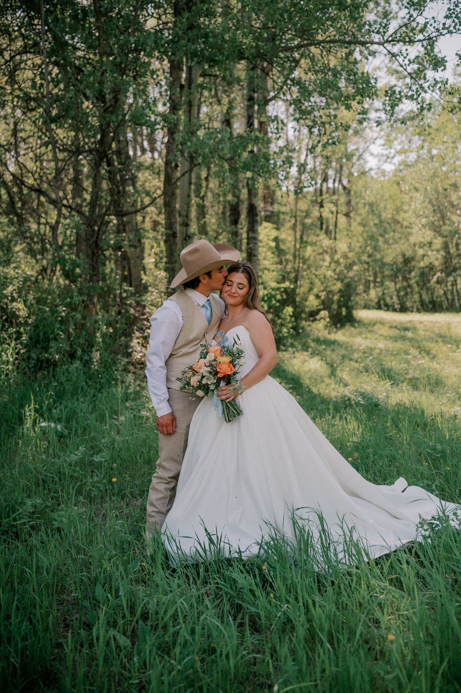 Bride and groom embracing in lush green forest, groom in cowboy hat, bride holding colorful bouquet.