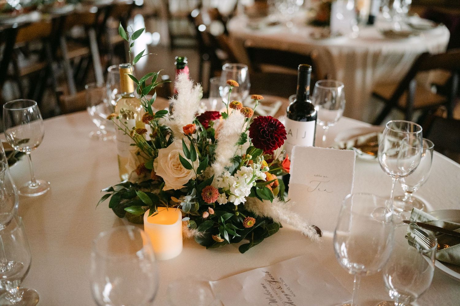 Elegant wedding reception table with lush floral centerpiece, wine bottle, candles, and glassware on white linen.