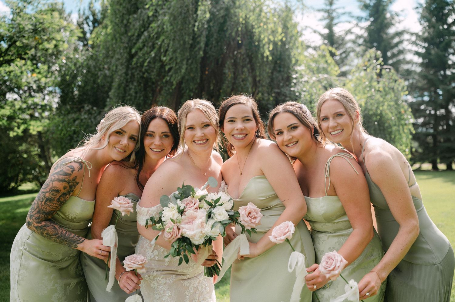 Bride in white gown holds pink bouquet surrounded by five bridesmaids in sage green dresses outdoors.