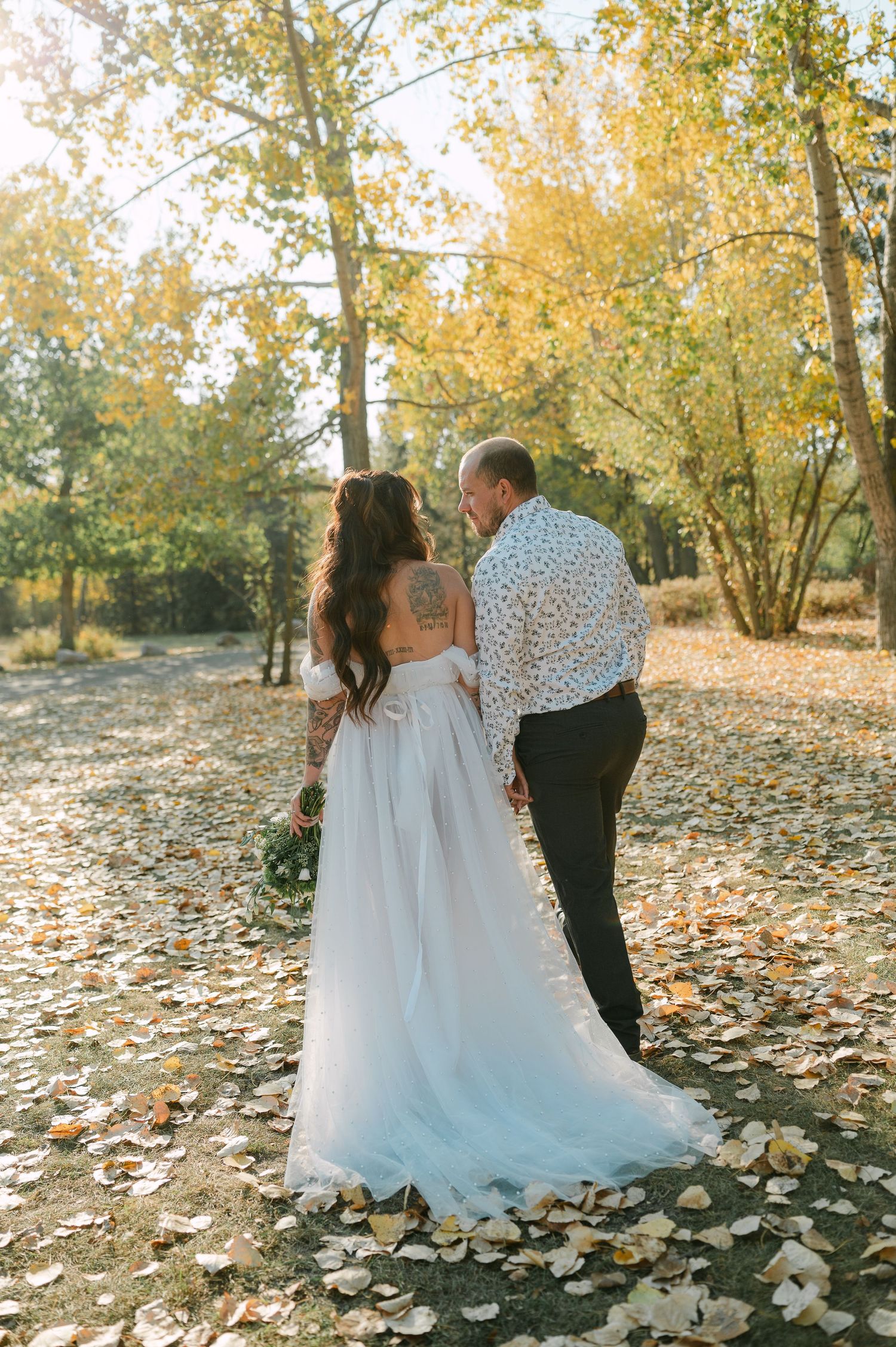 Bride and groom walking hand-in-hand through autumn forest with golden fallen leaves on the ground.
