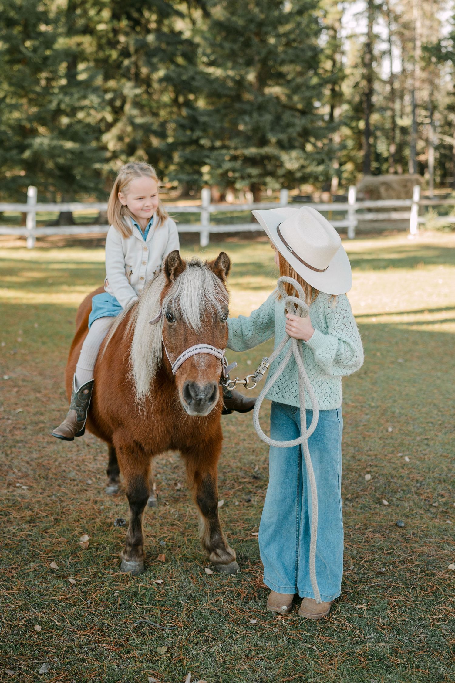 Young girl riding a brown pony led by her sister in a cowboy hat in a fenced pasture. Alberta heritage photoshoot