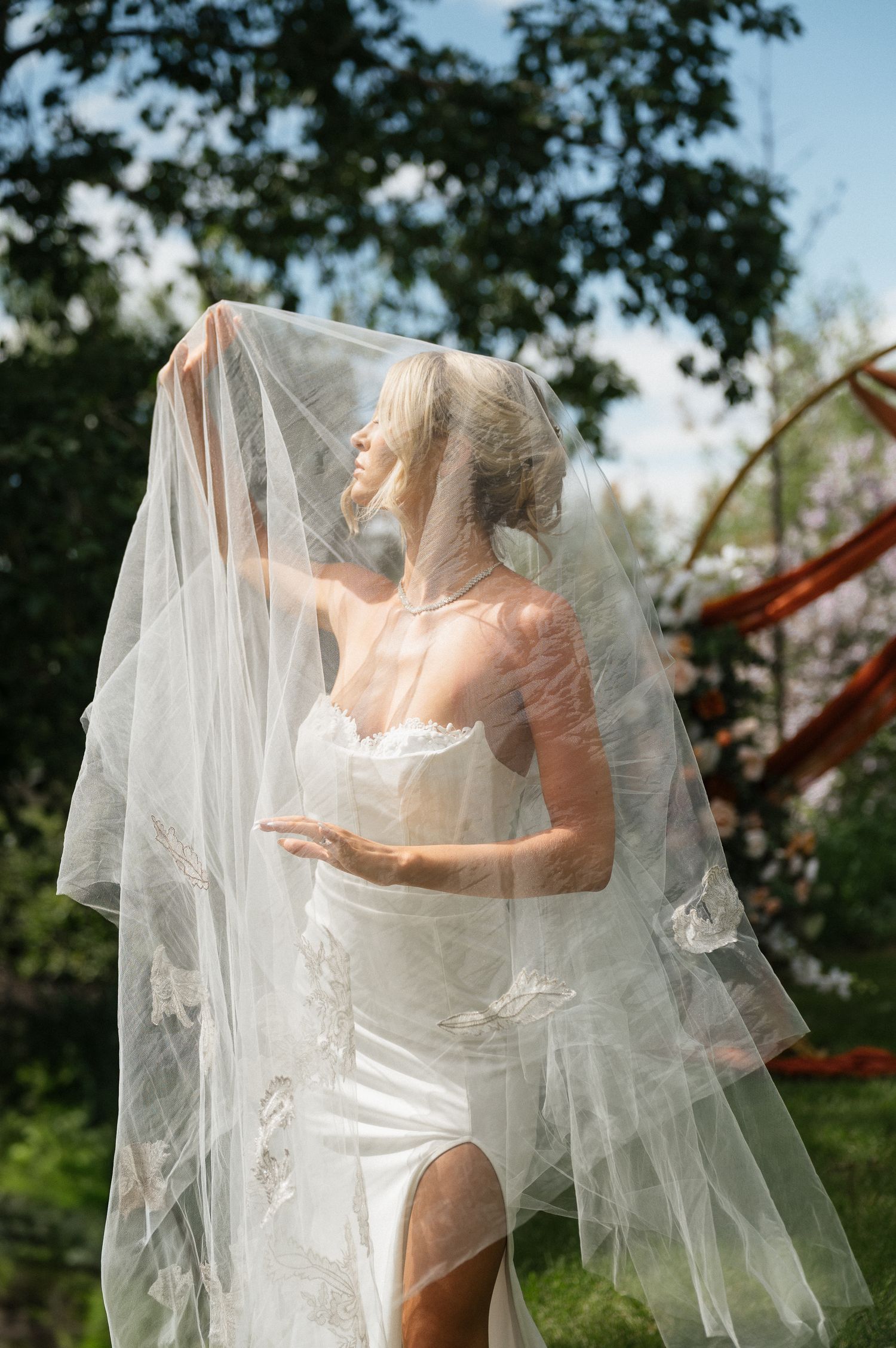 Blonde bride in a fitted white gown lifting her lace veil outdoors in a sunlit garden setting.