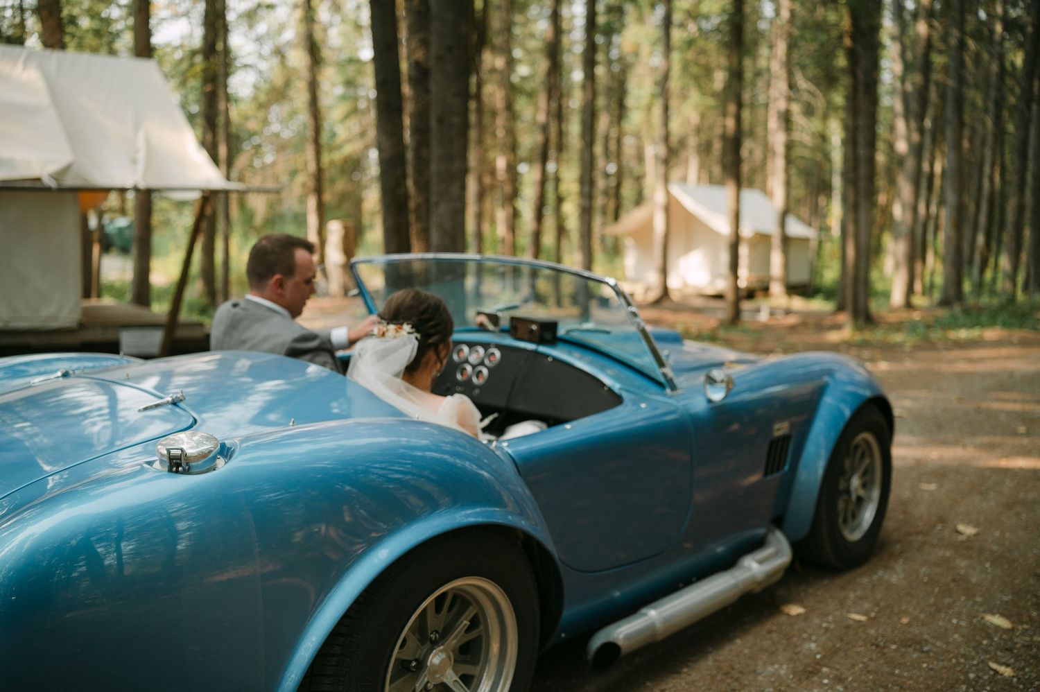 Bride and groom riding in a classic blue vintage convertible sports car through a wooded forest setting.