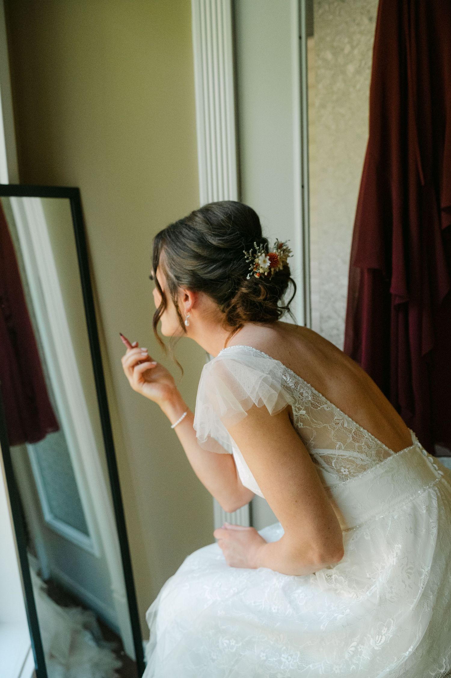 Bride in lace wedding gown applying makeup while seated beside a mirror with floral hair accessory.