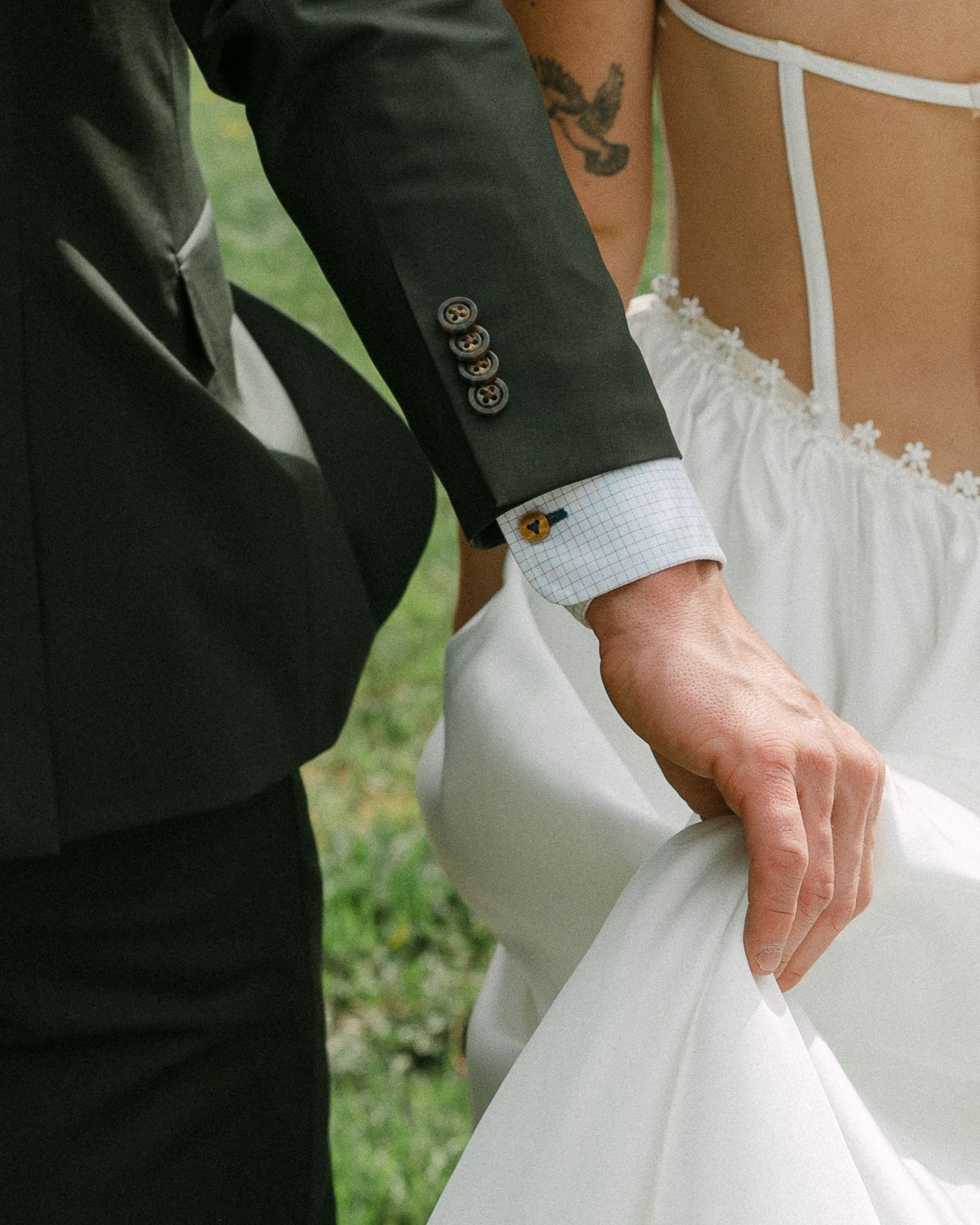 Close-up of groom in dark suit holding bride's white wedding dress, tattoo visible on bride's back.