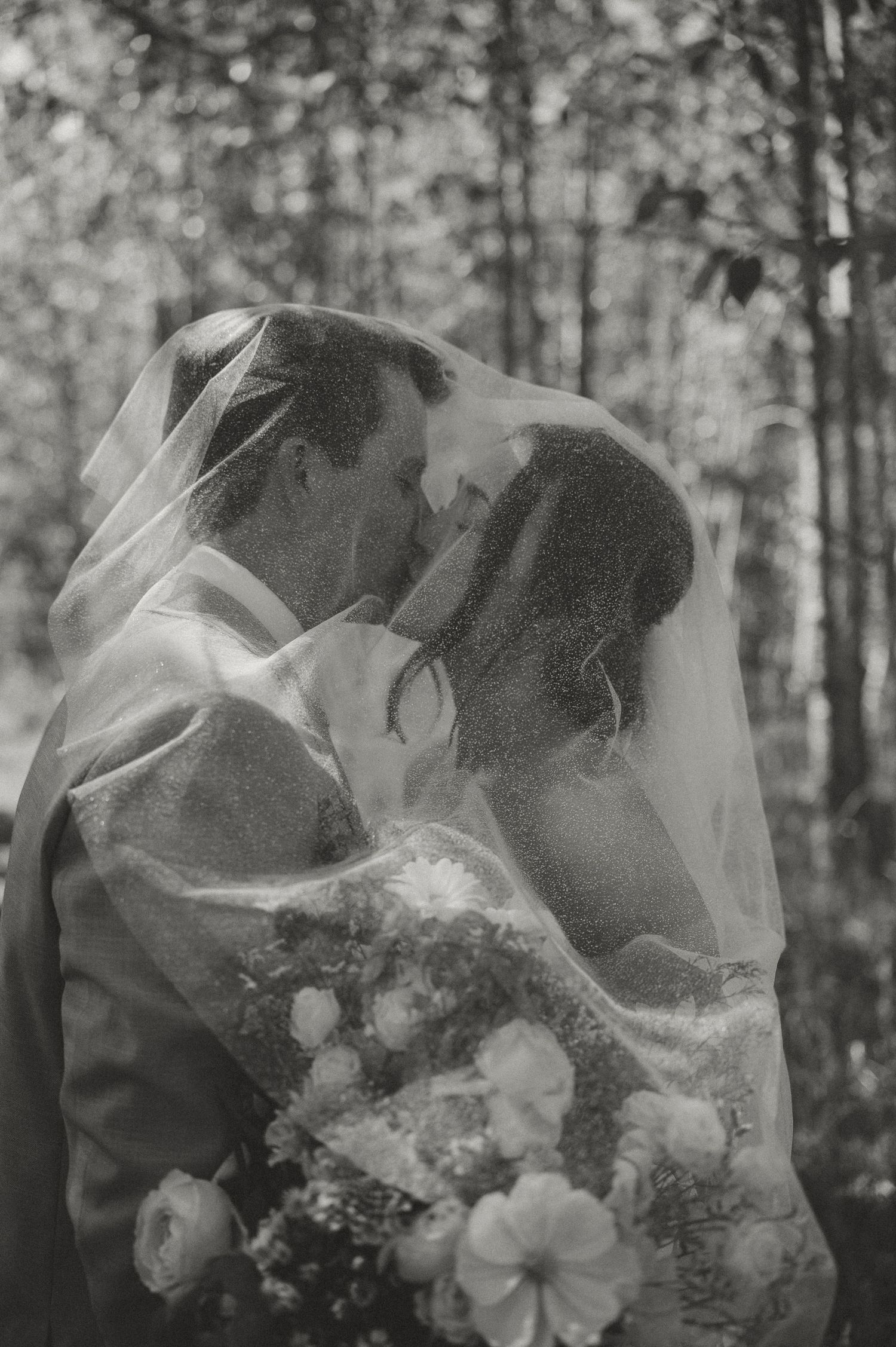 Black and white romantic photo of bride and groom kissing under a wedding veil in a forest.
