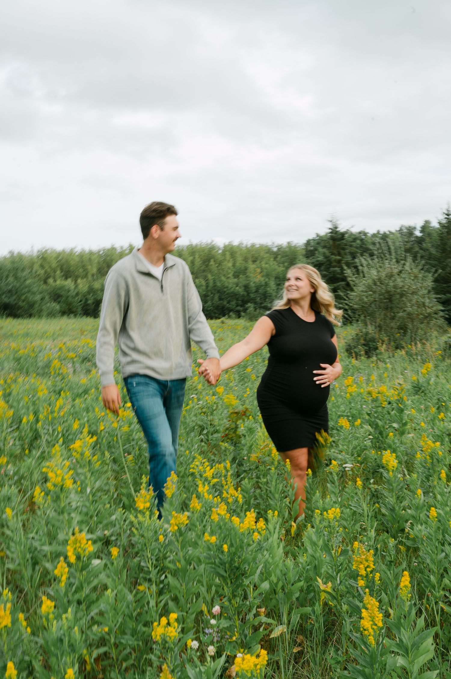 Pregnant couple holding hands walking through a field of yellow wildflowers, woman in black dress cradling baby bump. Materinty Session