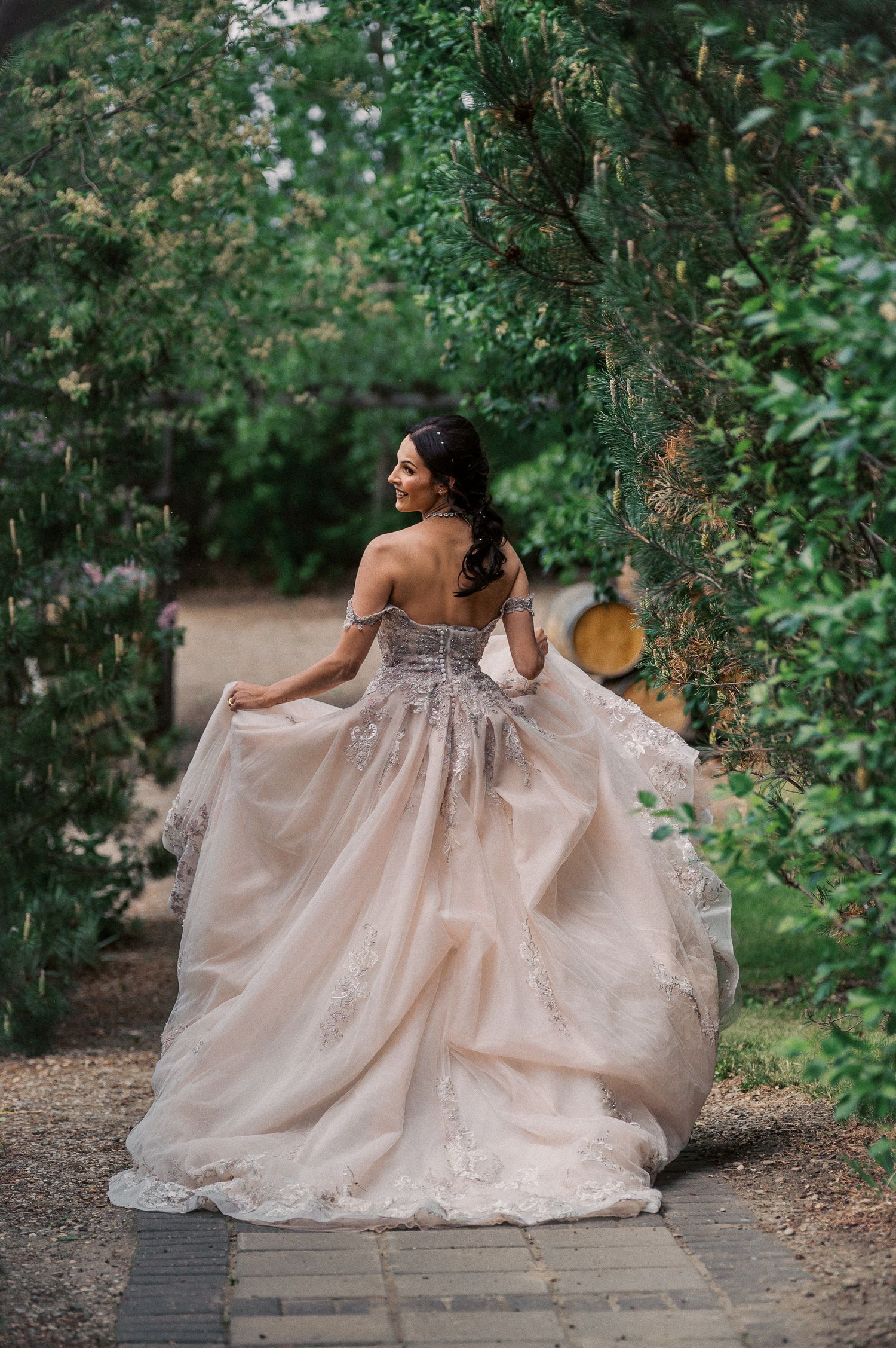 Bride in a stunning blush off-shoulder ballgown twirling under a green garden archway.