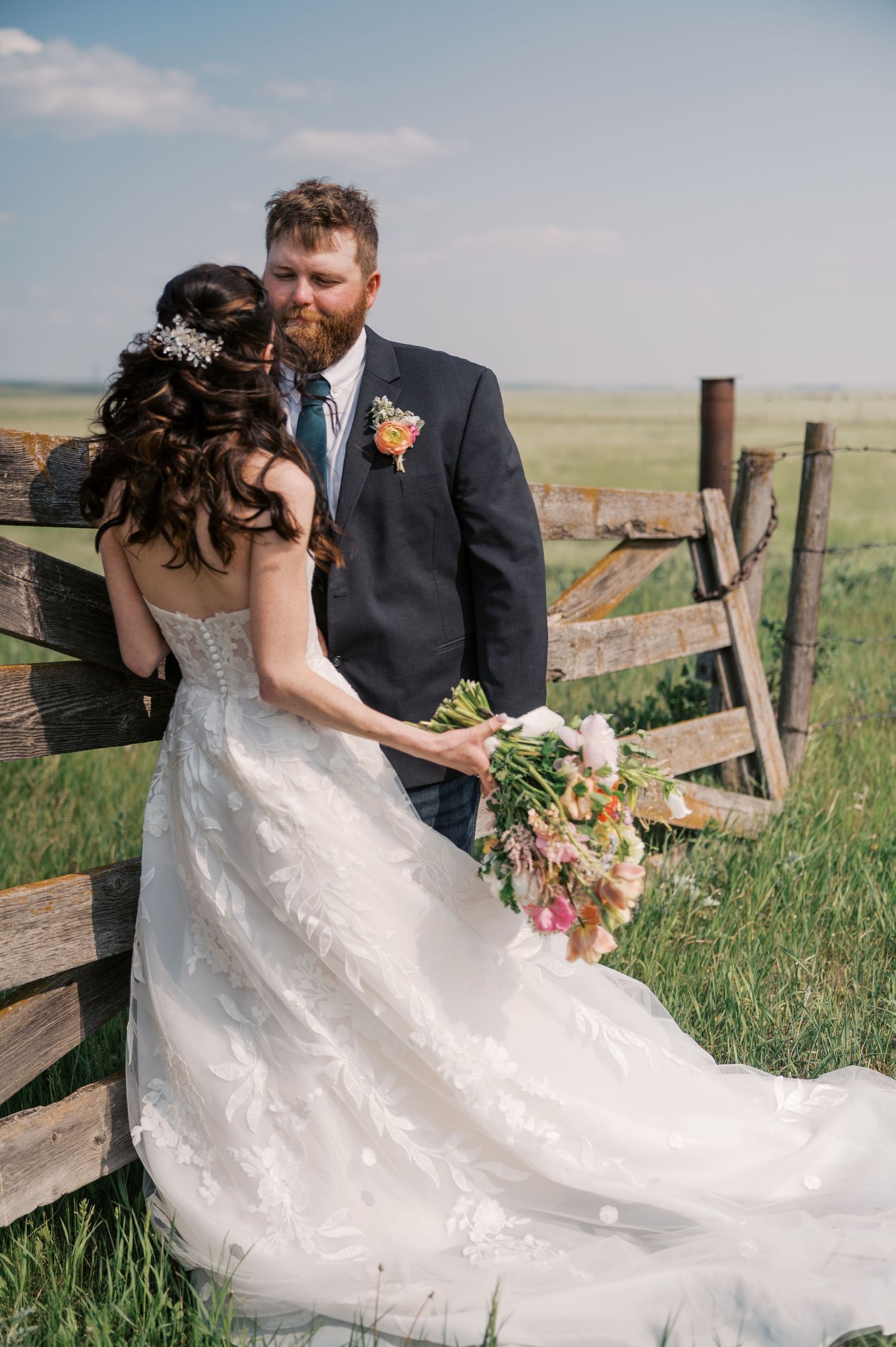 Bride in lace gown holding colorful bouquet embraces bearded groom in navy suit by rustic wooden fence in green field.