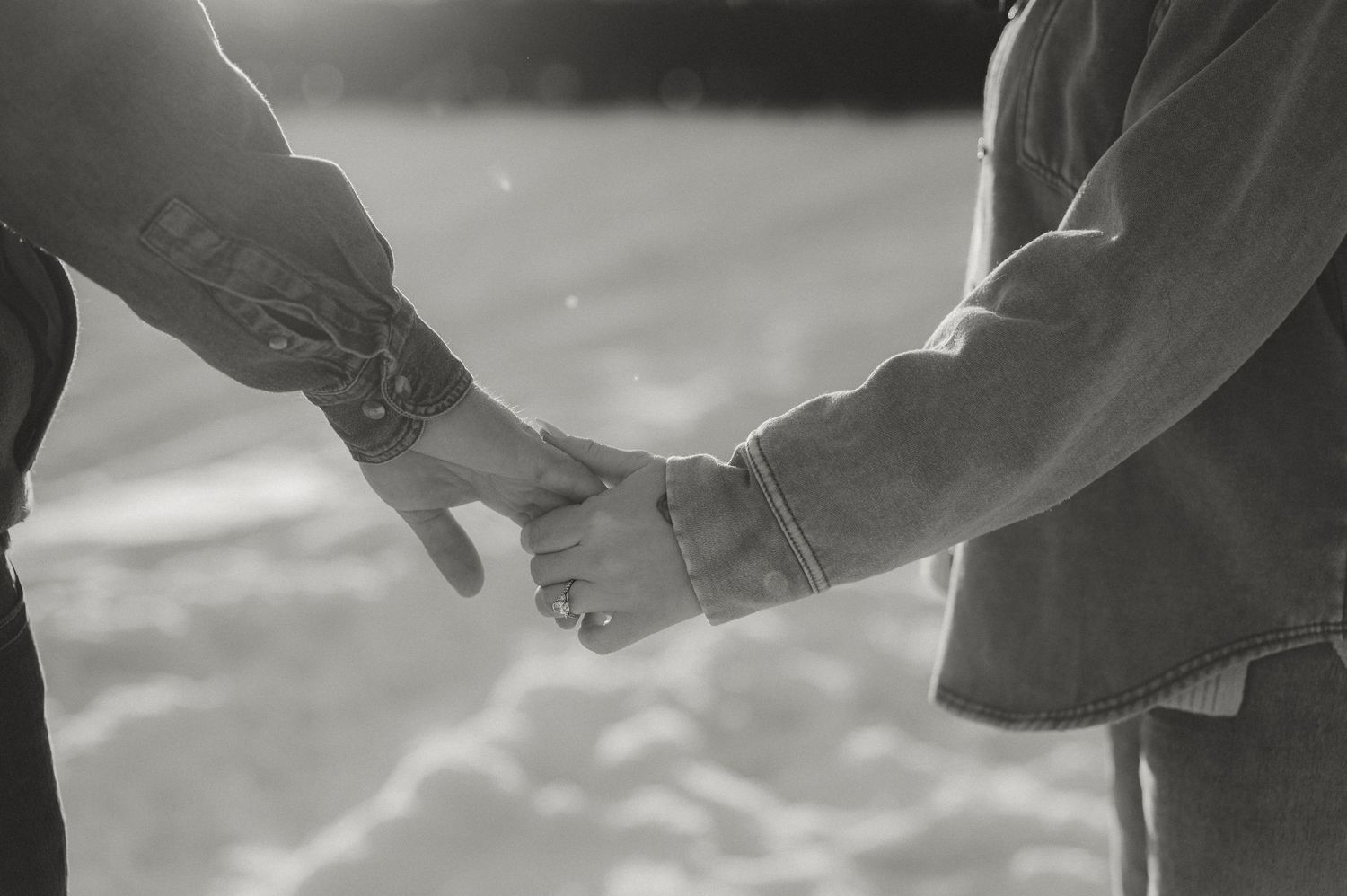 Black and white romantic photo of couple holding hands tenderly in snowy winter landscape.
