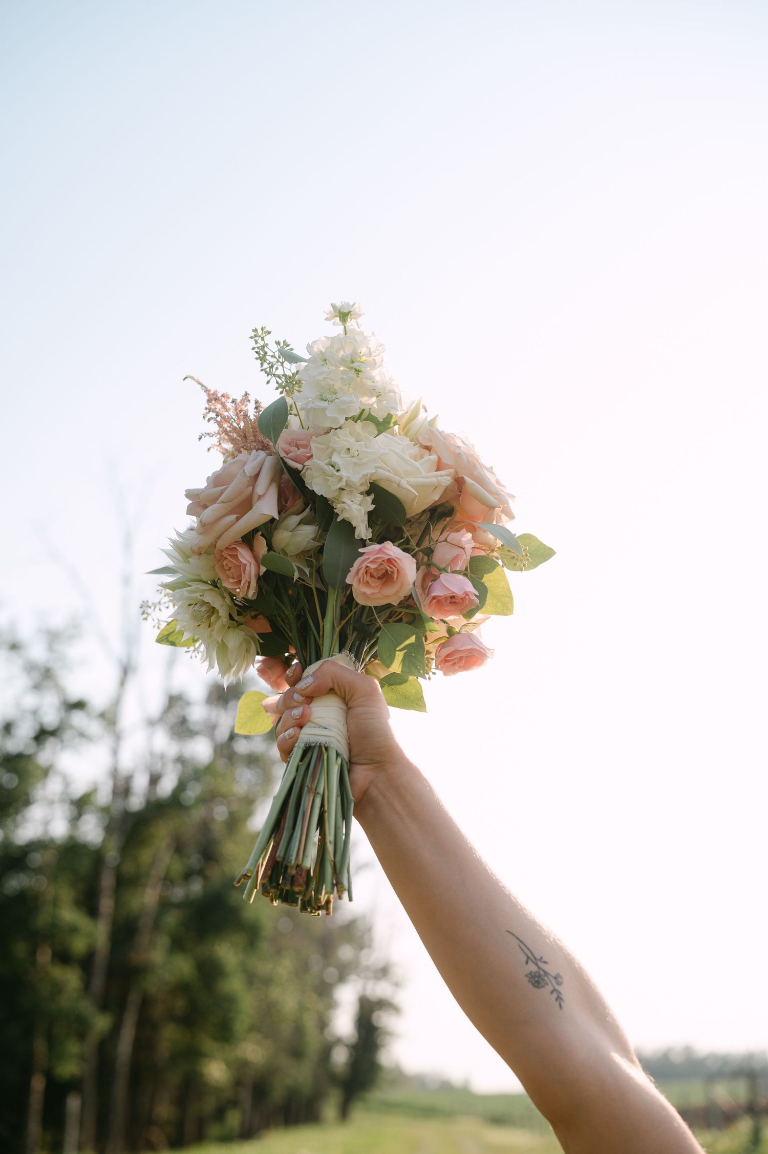 A tattooed hand holds a bridal bouquet of pink and white roses aloft against a bright sky.