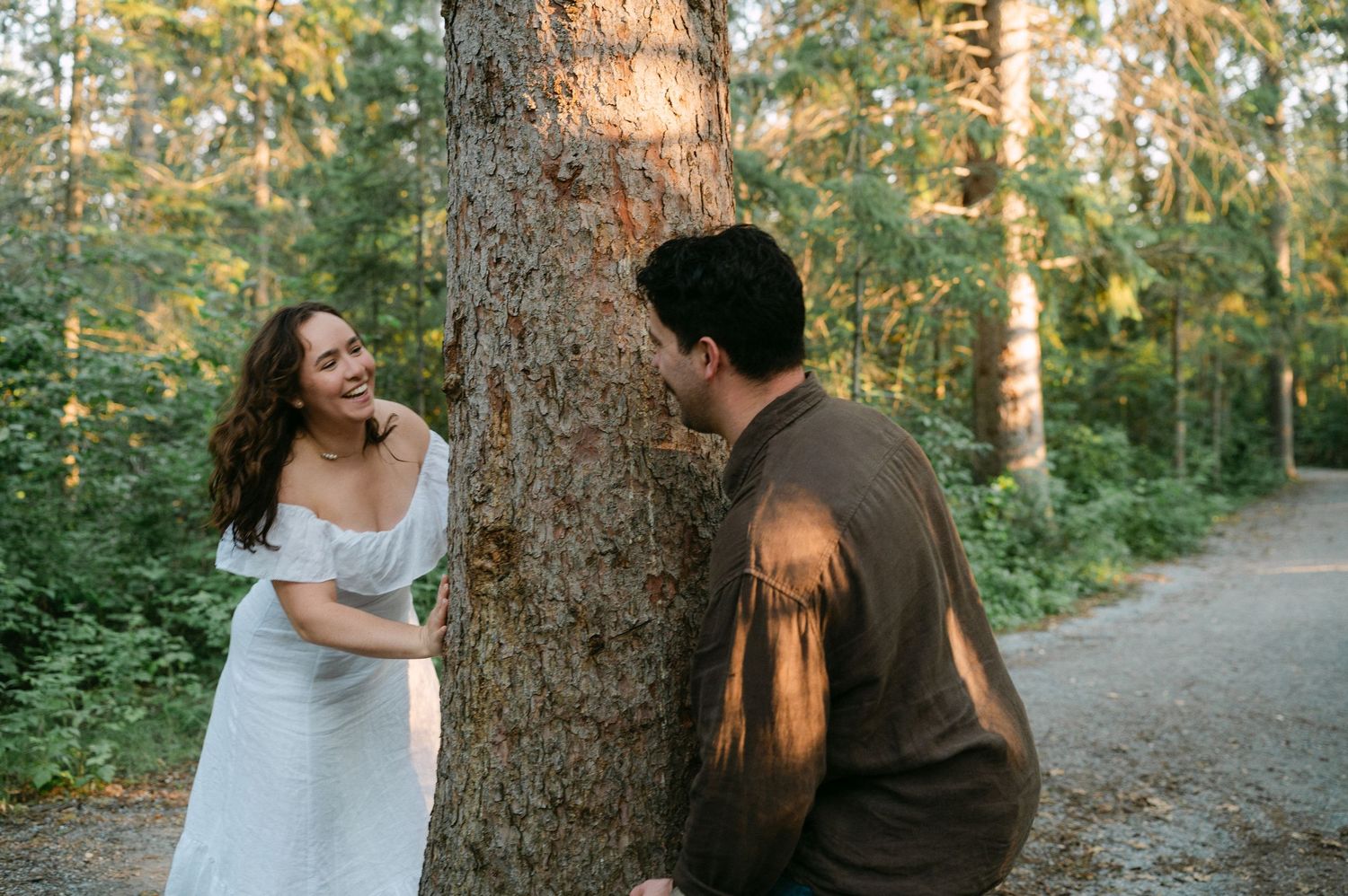 Couple laughing together by a tree in a sunlit forest, woman in white dress, man in brown sweater.