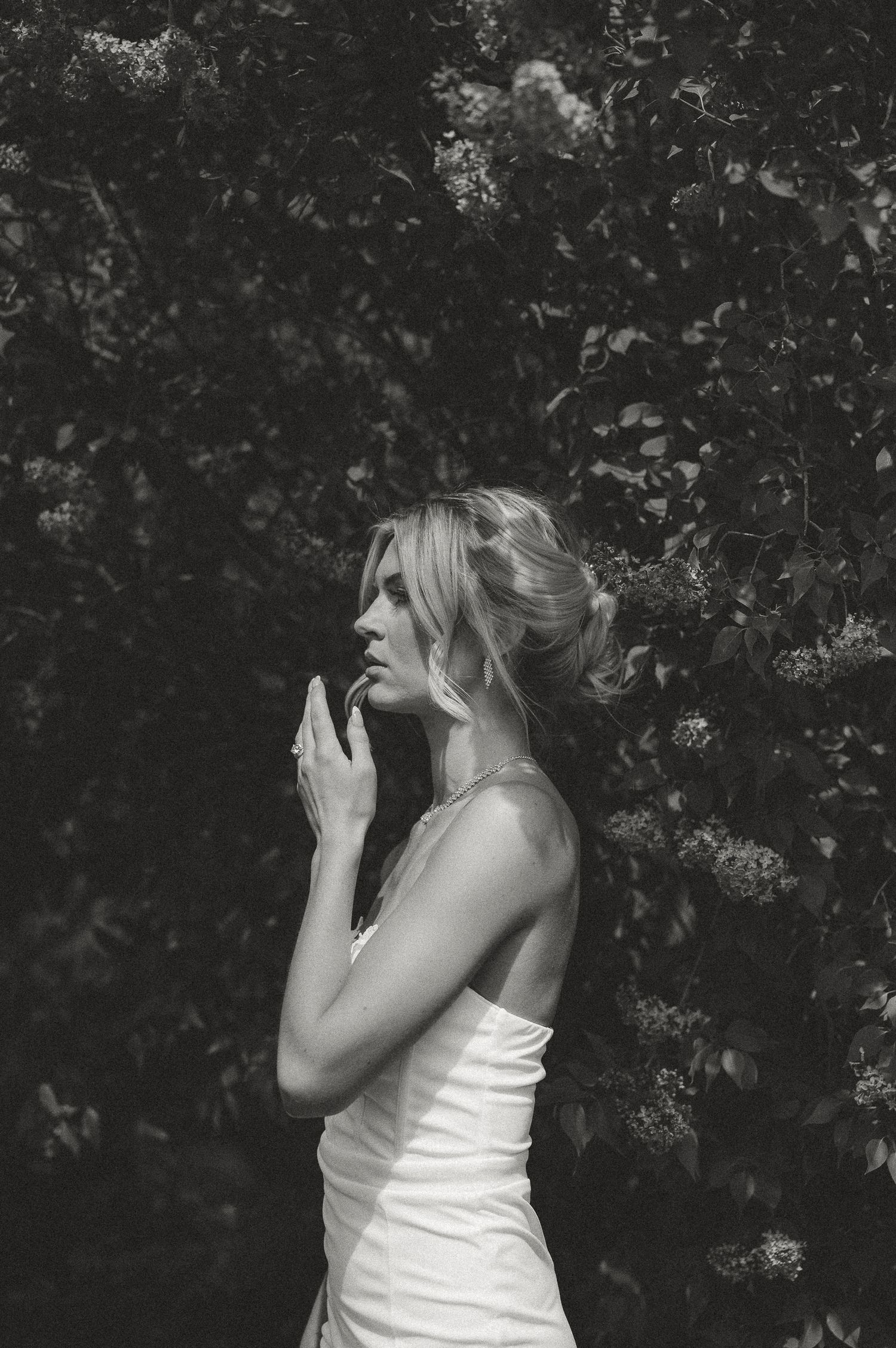 Black and white portrait of a bride in a white gown standing before lush green foliage.