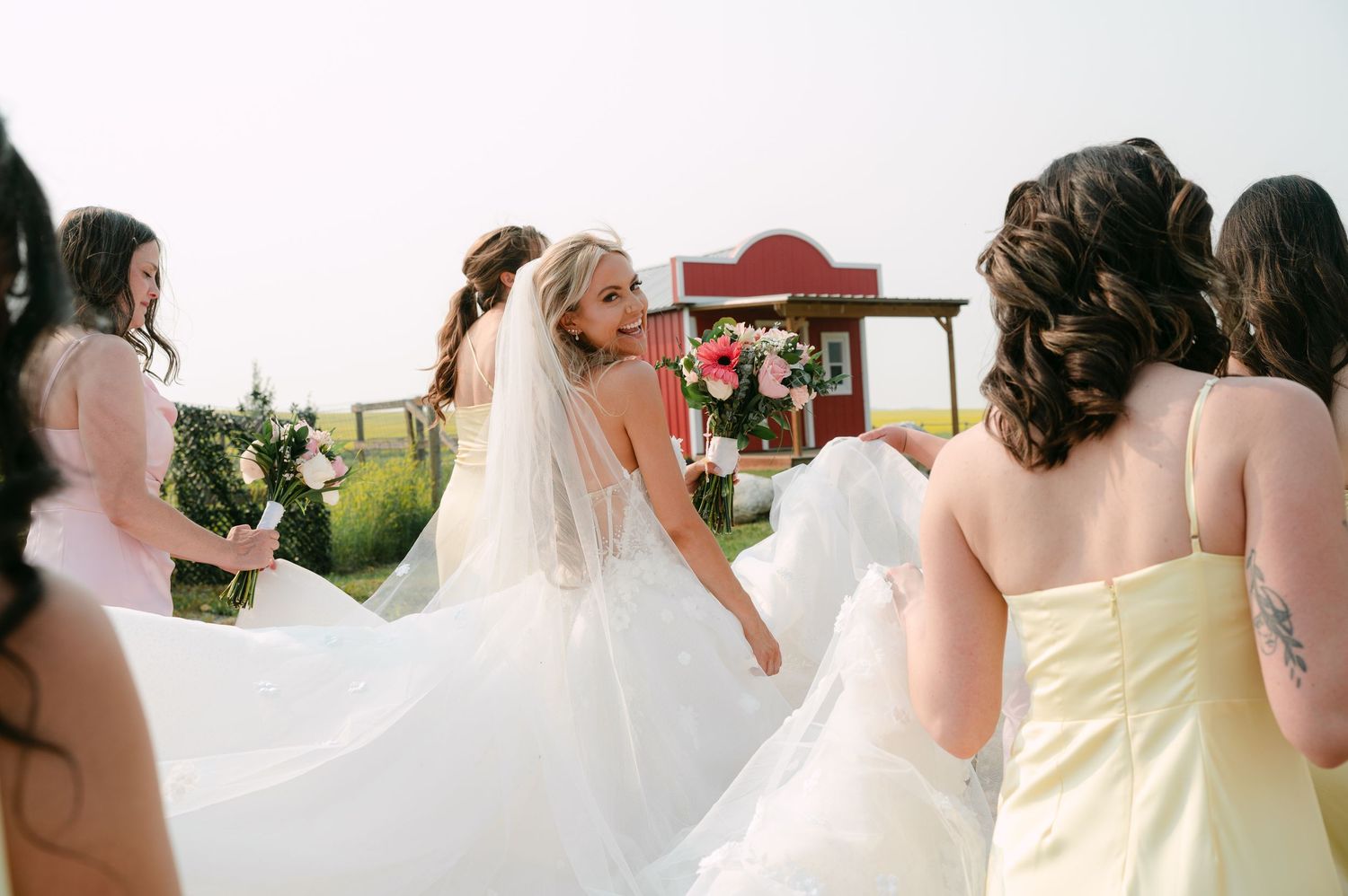 Bride in white gown and veil surrounded by bridesmaids in pink and yellow dresses at outdoor farm wedding.