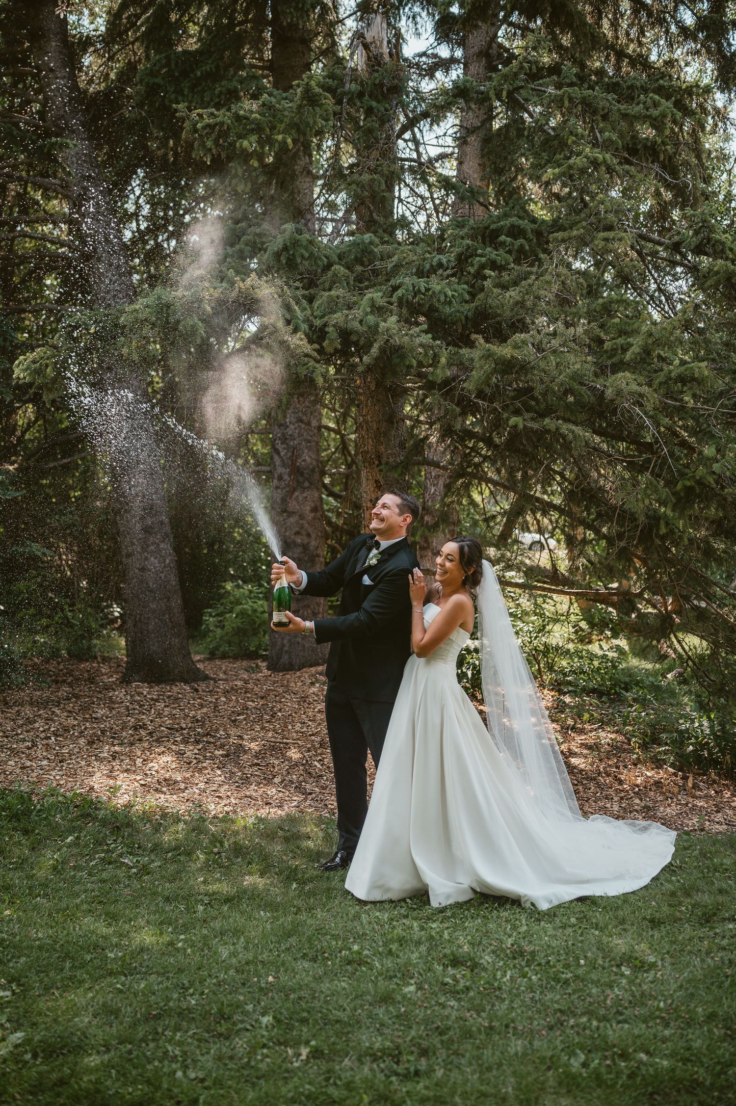 Newlyweds celebrate with champagne spray outdoors among tall evergreen trees on their wedding day.