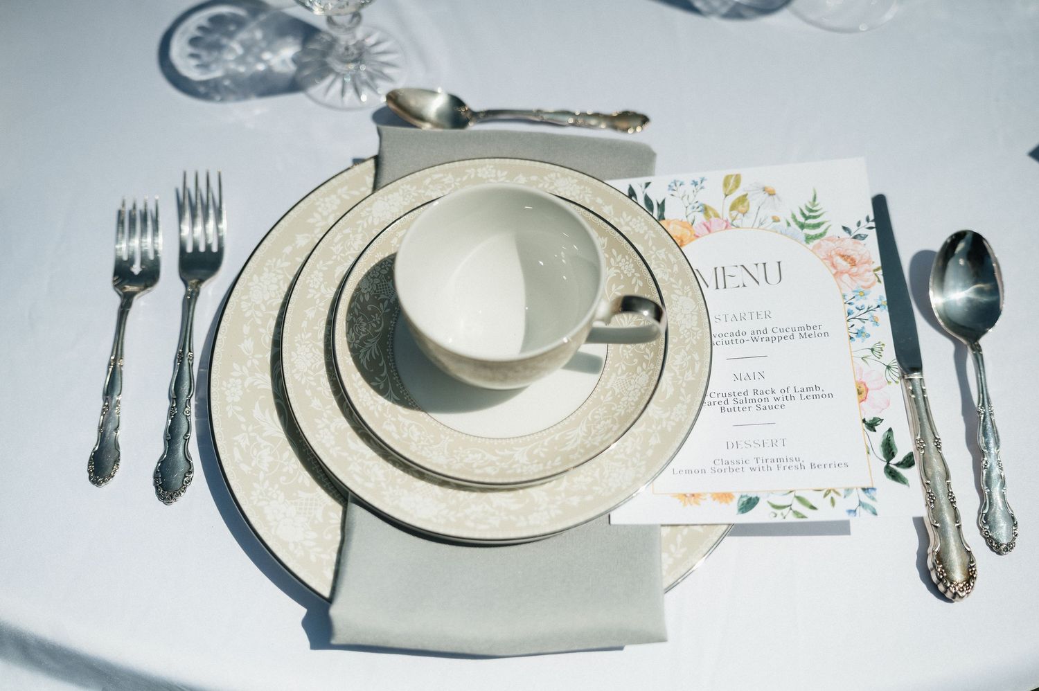 Elegant wedding place setting with silver-rimmed china, teacup, silverware, and floral menu card.