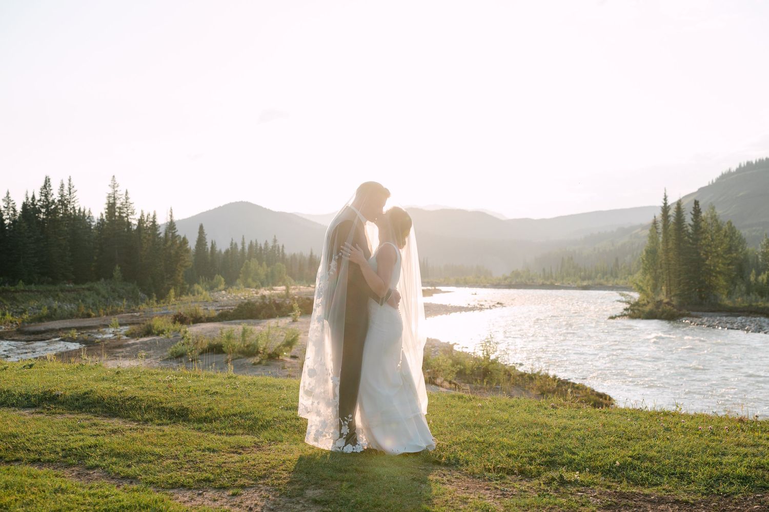 Bride and groom share a romantic kiss at sunset by a mountain river, with evergreen trees and peaks in the background.