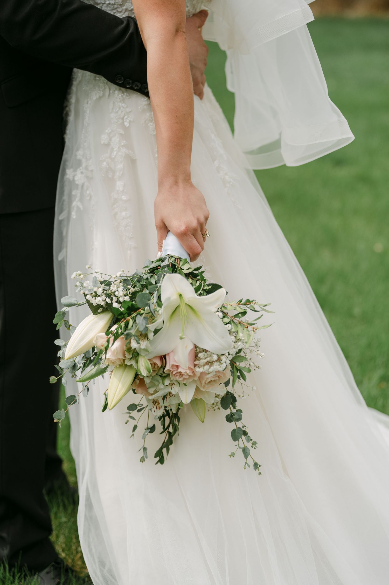 Close-up of a bride holding a white floral bouquet with lilies and eucalyptus beside her lace wedding gown.